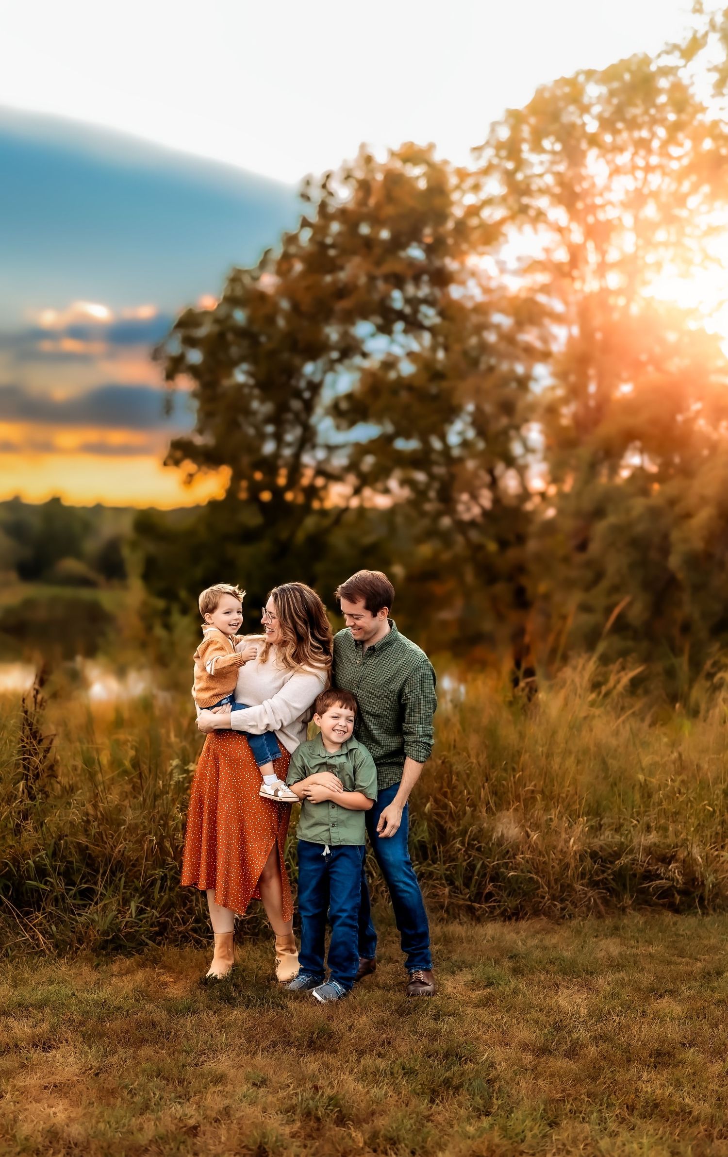 Family of four poses at golden hour sunset near a pond, parents holding children in an autumn outdoor portrait.