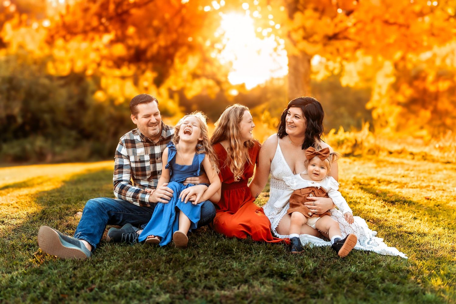 Family of five laughing together on grass surrounded by vibrant autumn foliage during golden hour.