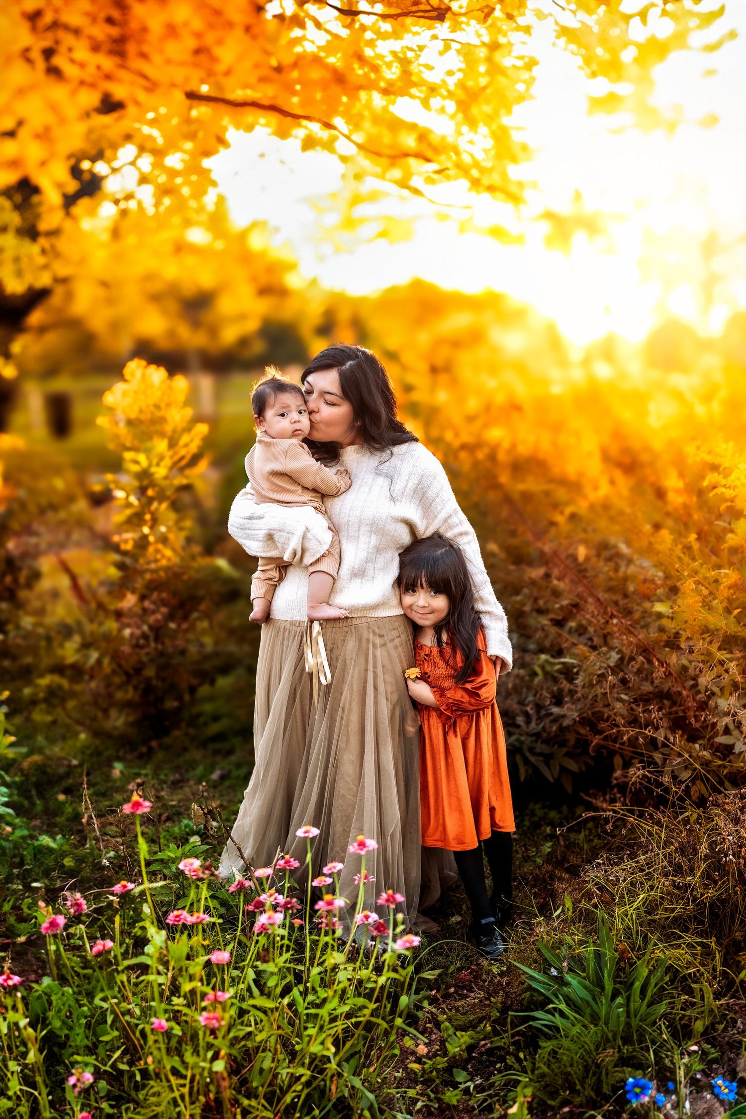 Mother kissing baby while daughter hugs her in a sunlit autumn garden with golden foliage and pink flowers.