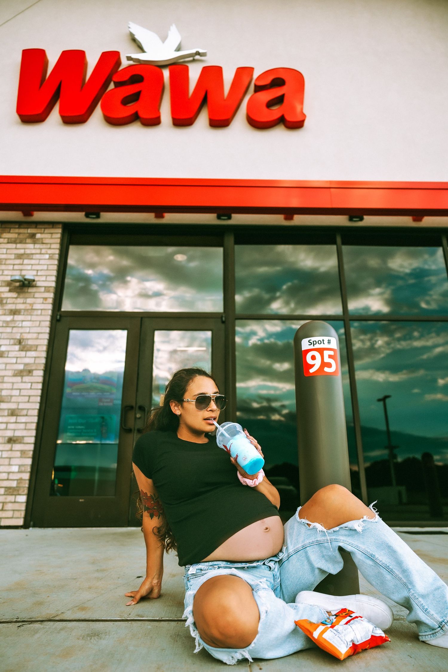 Pregnant woman in sunglasses sits outside Wawa store drinking a beverage, wearing ripped jeans and black top.