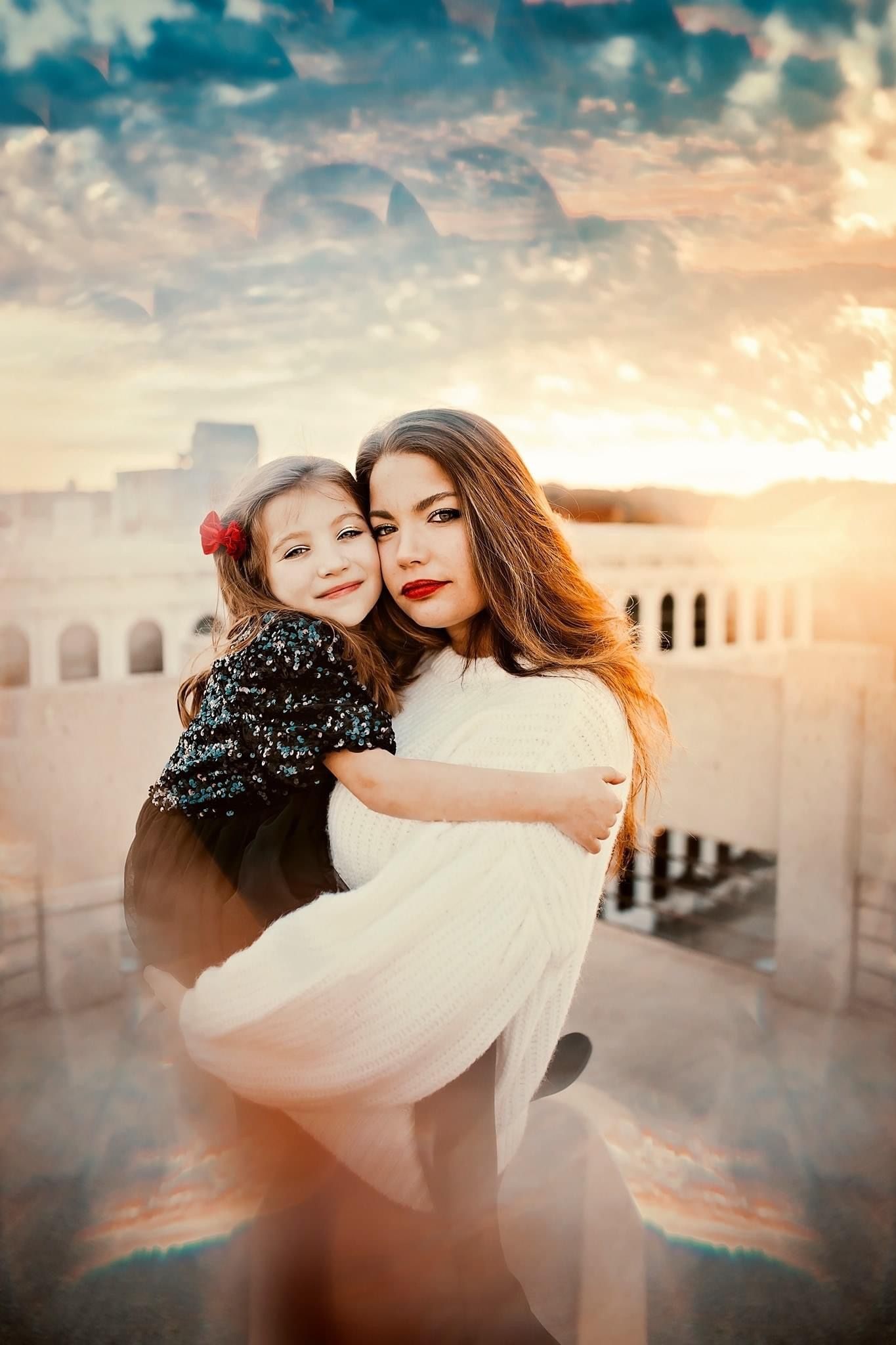 A woman in white holds a young girl in a black dress, hugging at sunset with arched architecture in the background.
