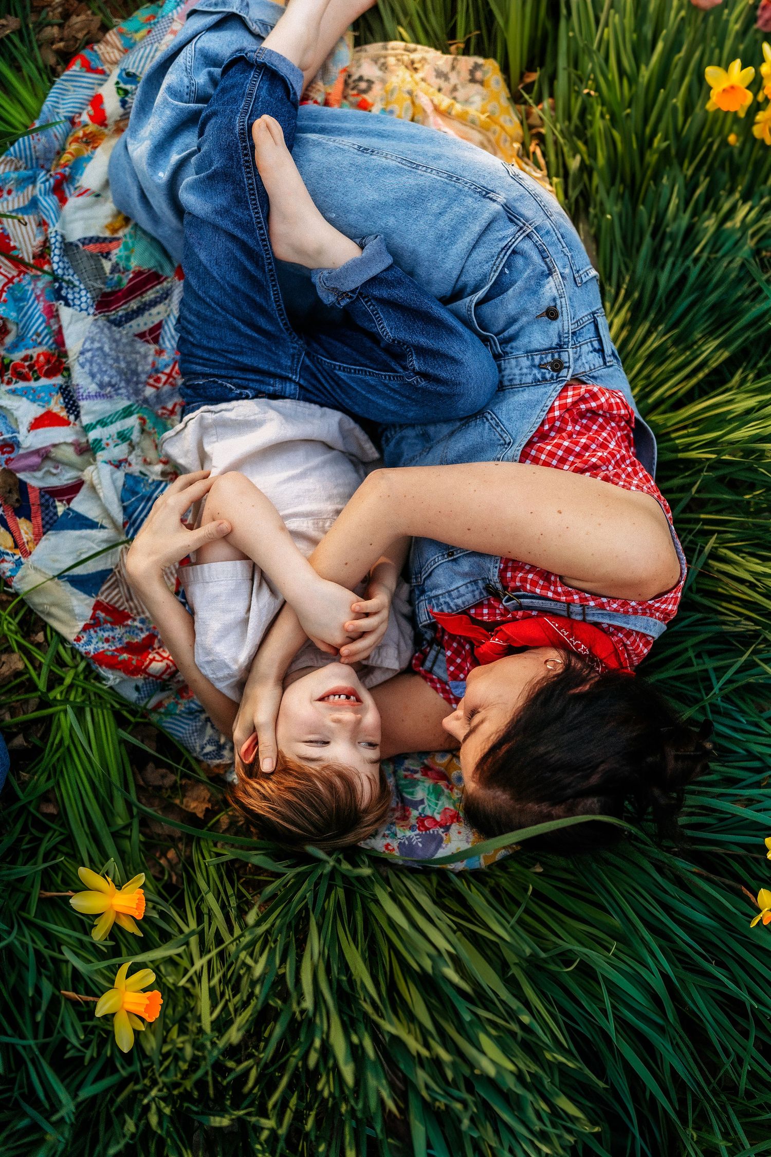 Mother and child lying on a colorful quilt blanket surrounded by green grass and yellow daffodils outdoors.