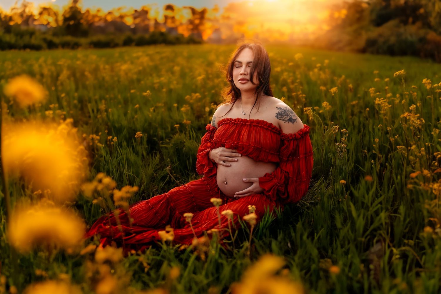 Pregnant woman in red dress sitting in a sunny field of yellow wildflowers during golden hour maternity photoshoot.