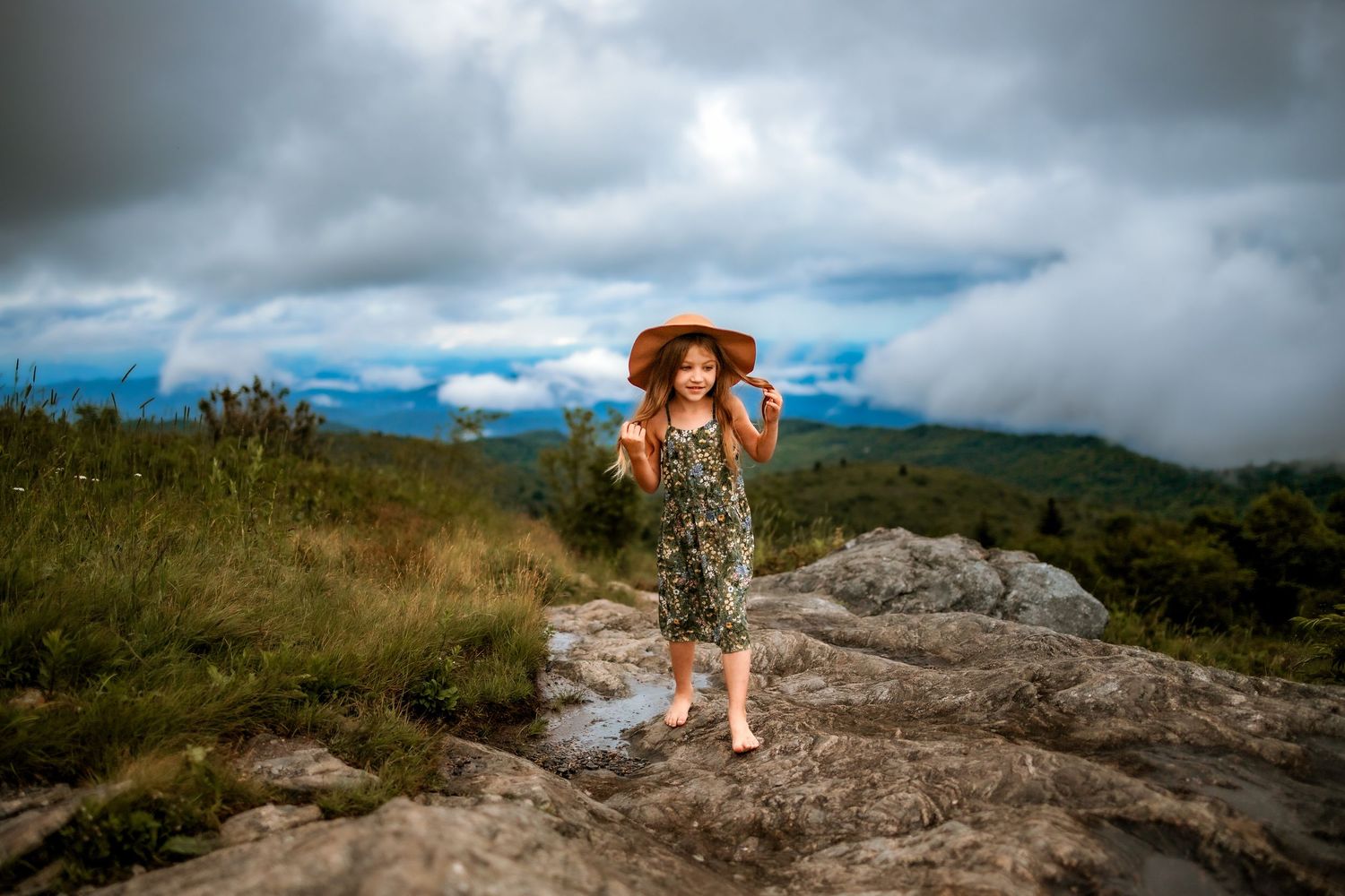 A young girl in a floral dress and sun hat runs barefoot on rocky terrain with mountains and cloudy sky behind her.