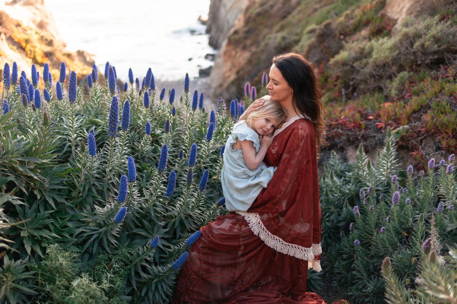 Mother in red dress holds young child among tall blue wildflowers on a coastal cliffside at golden hour.