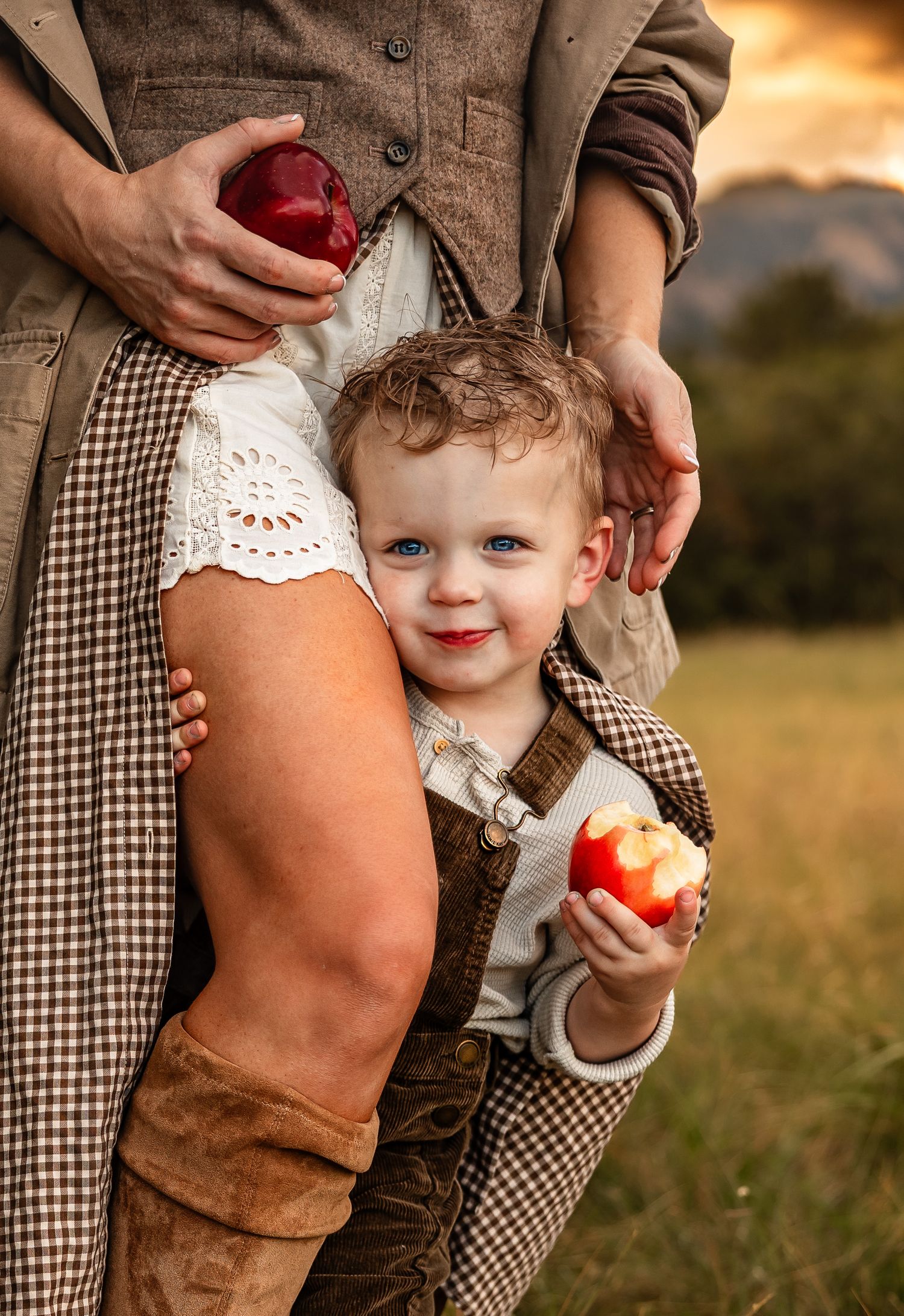 Smiling toddler boy in overalls holds a bitten red apple while standing beside a woman in a plaid dress and boots in a golden autumn field.