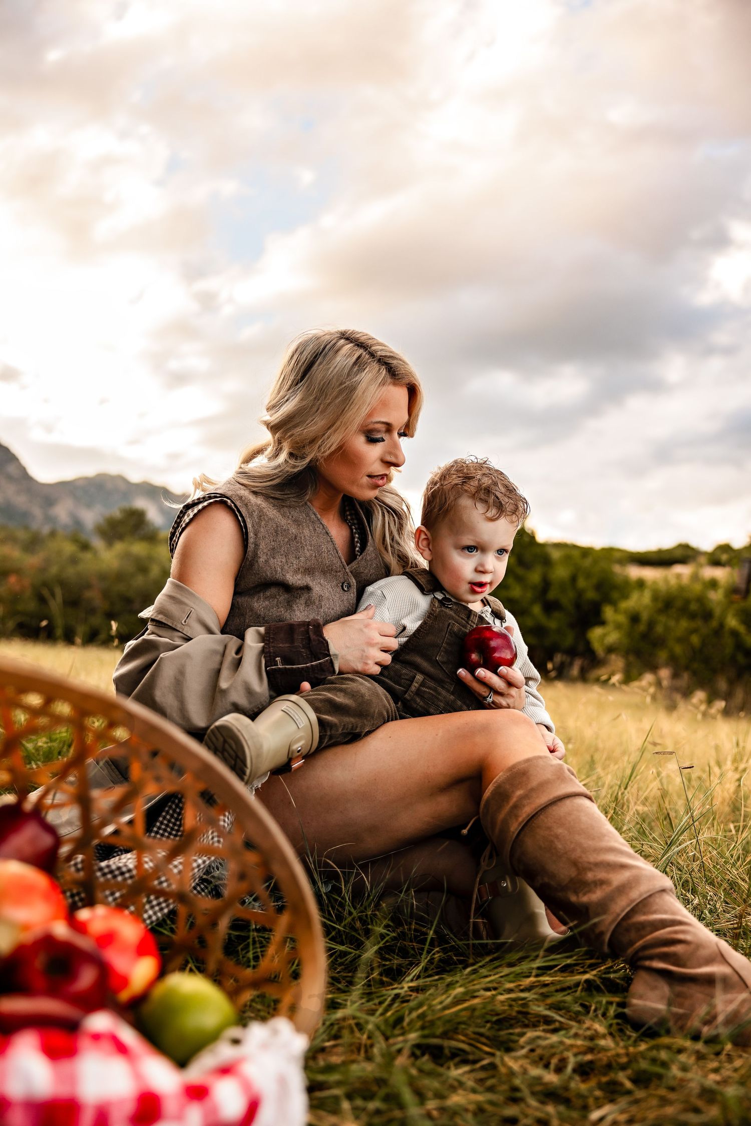 Blonde mother in a vest and tall suede boots sits on grass holding her toddler son and a red apple beside a basket of fruit at sunset.