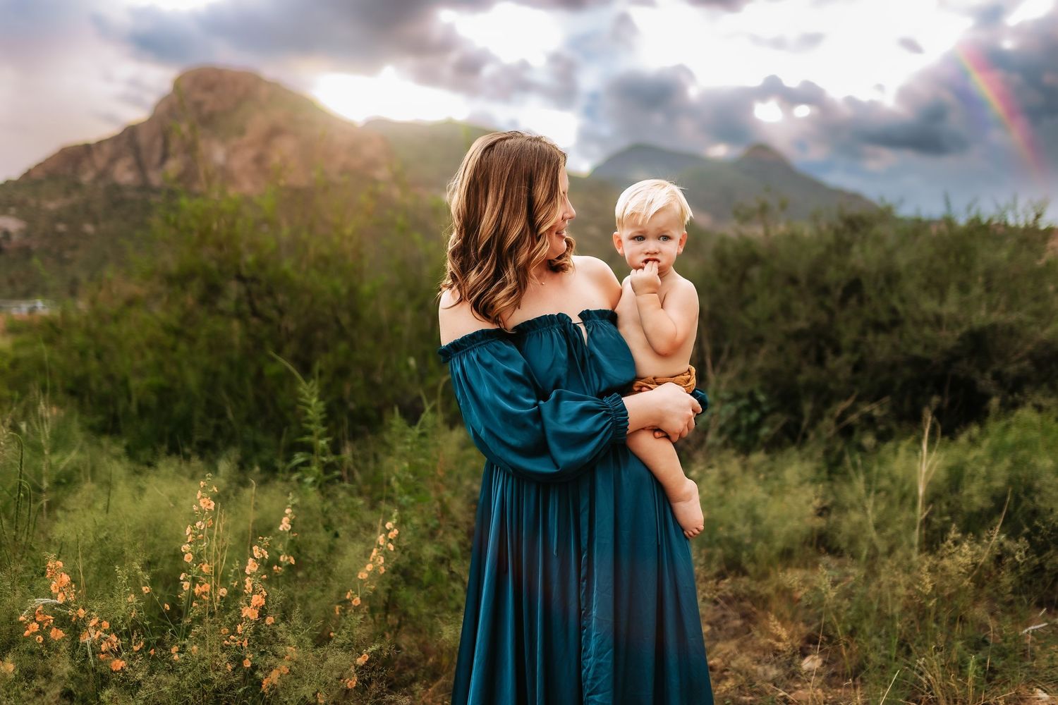 Mother in teal off-shoulder gown holds toddler boy outdoors with mountain and rainbow backdrop.