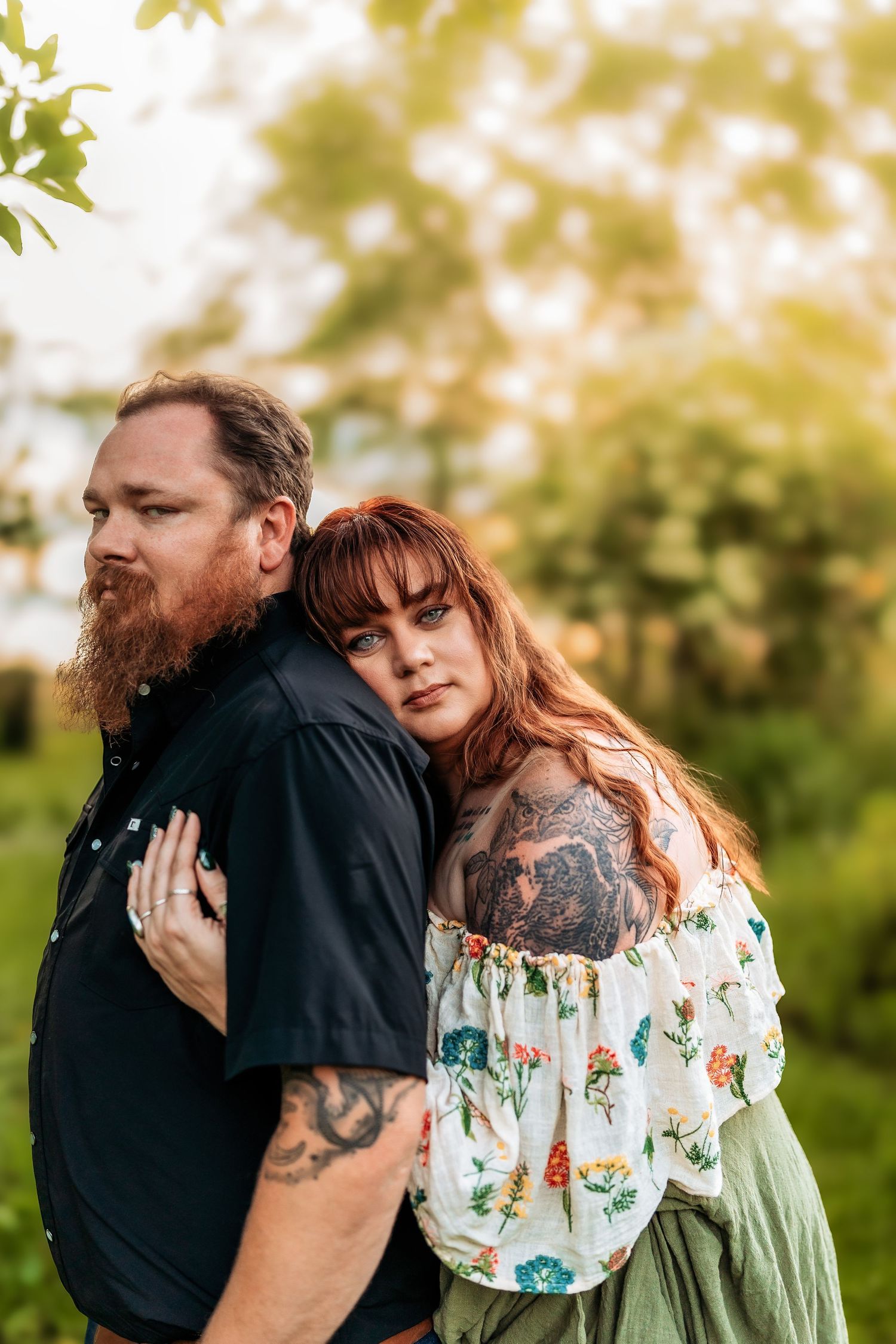 A tattooed couple poses outdoors, woman in floral top leans on bearded man in black shirt, surrounded by lush greenery.