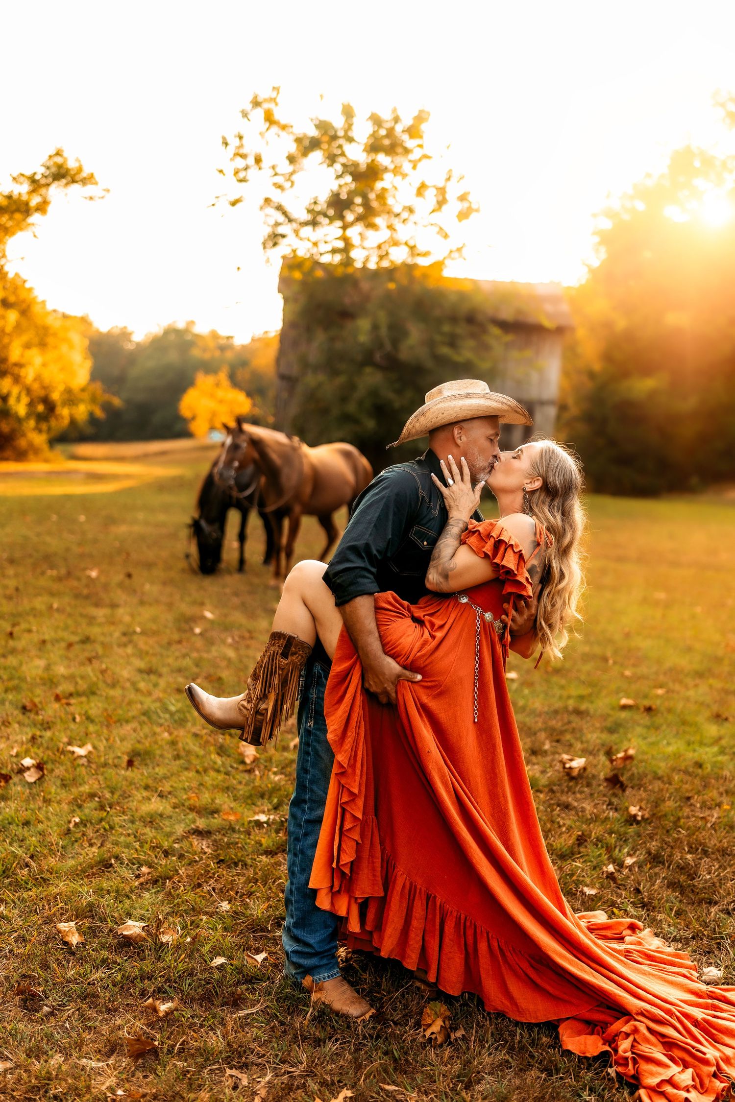 A couple shares a romantic kiss at golden hour, man in cowboy hat lifting woman in orange dress, horse grazing in background.