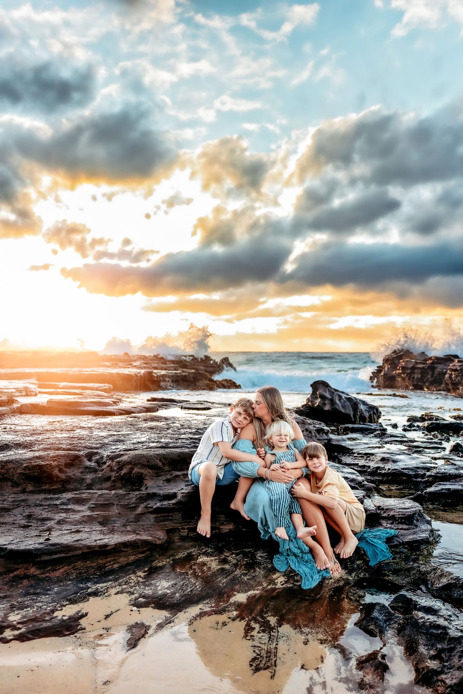 Family of four sitting on rocky shore at sunset with dramatic cloudy sky and ocean waves in background.