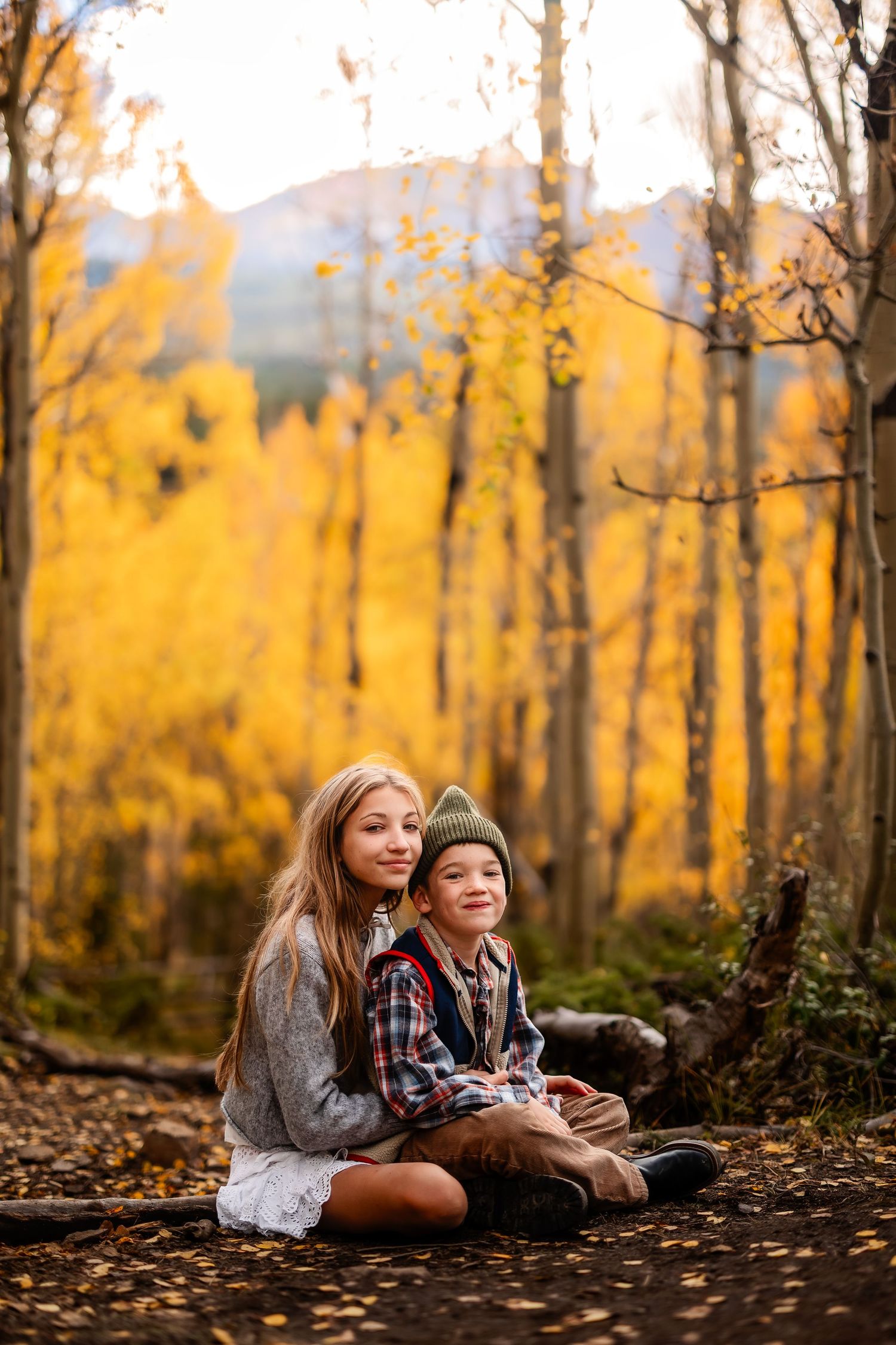 Two children sit together on a forest path surrounded by vibrant golden autumn aspen trees with mountains behind.