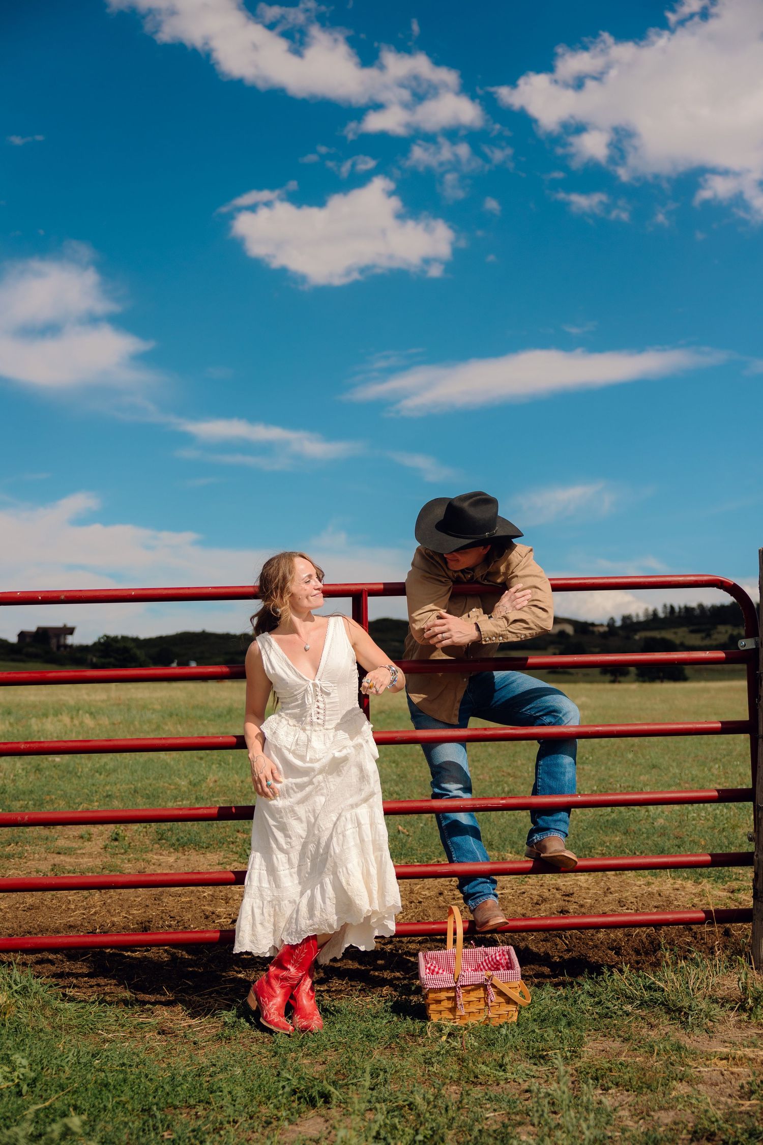 Couple in western attire by red farm gate, woman in white dress and red boots, man in cowboy hat, blue sky backdrop.