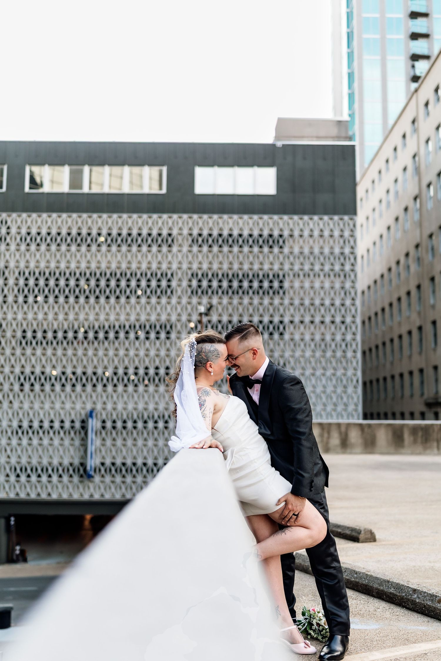 Couple sharing a romantic kiss in wedding attire against an urban building backdrop on a city rooftop.