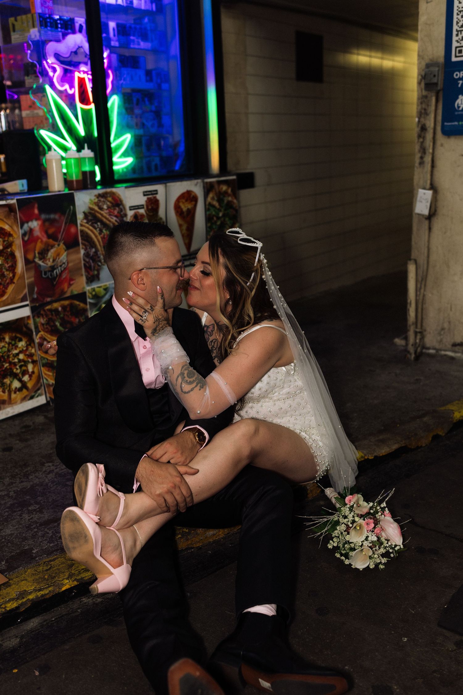 Newlywed couple kissing outside a neon-lit storefront, bride in white dress with veil holding bouquet.