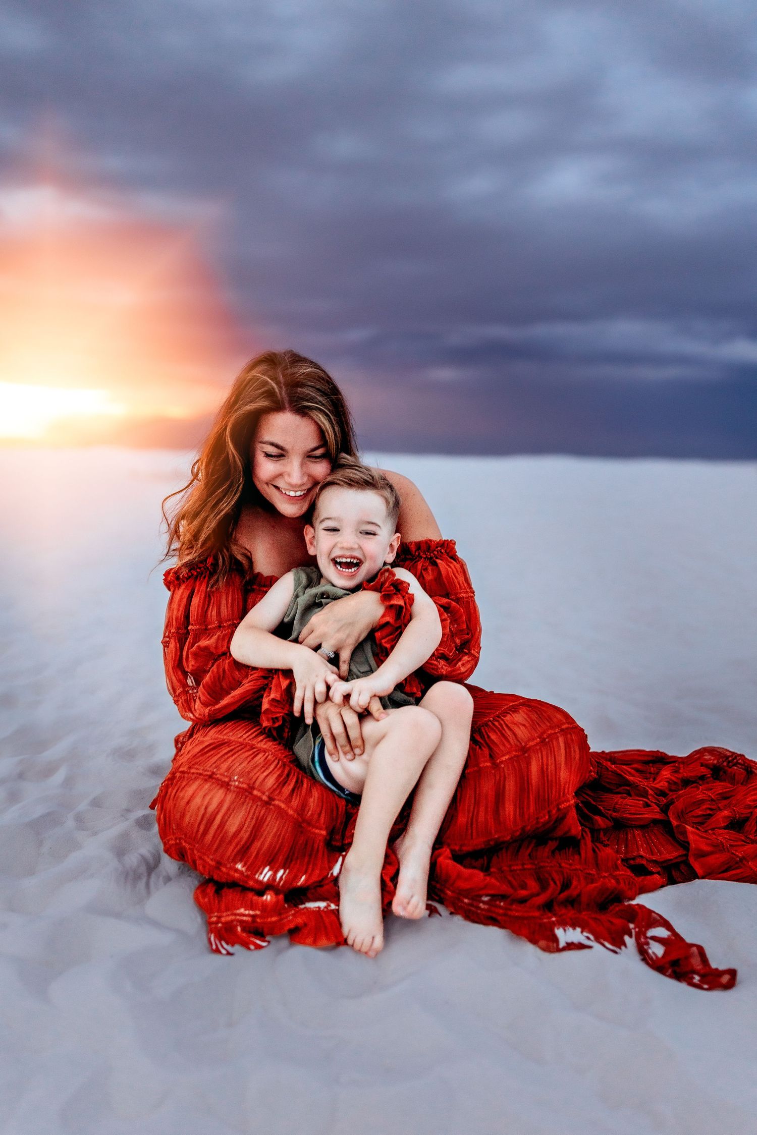 Woman in red dress hugging laughing child on white sand dunes at sunset with dramatic stormy sky.