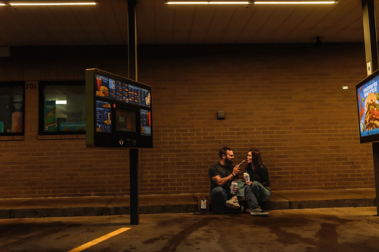 Couple sitting together against a brick wall at night near illuminated signs in a parking lot.