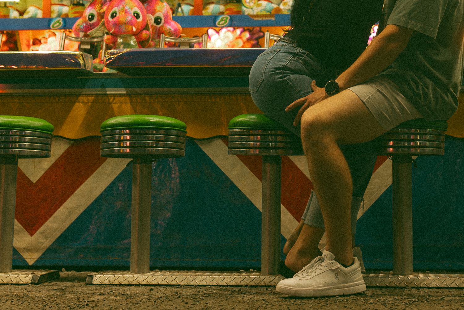Two people sit on green stools at a colorful carnival game booth at night, wearing shorts and sneakers.
