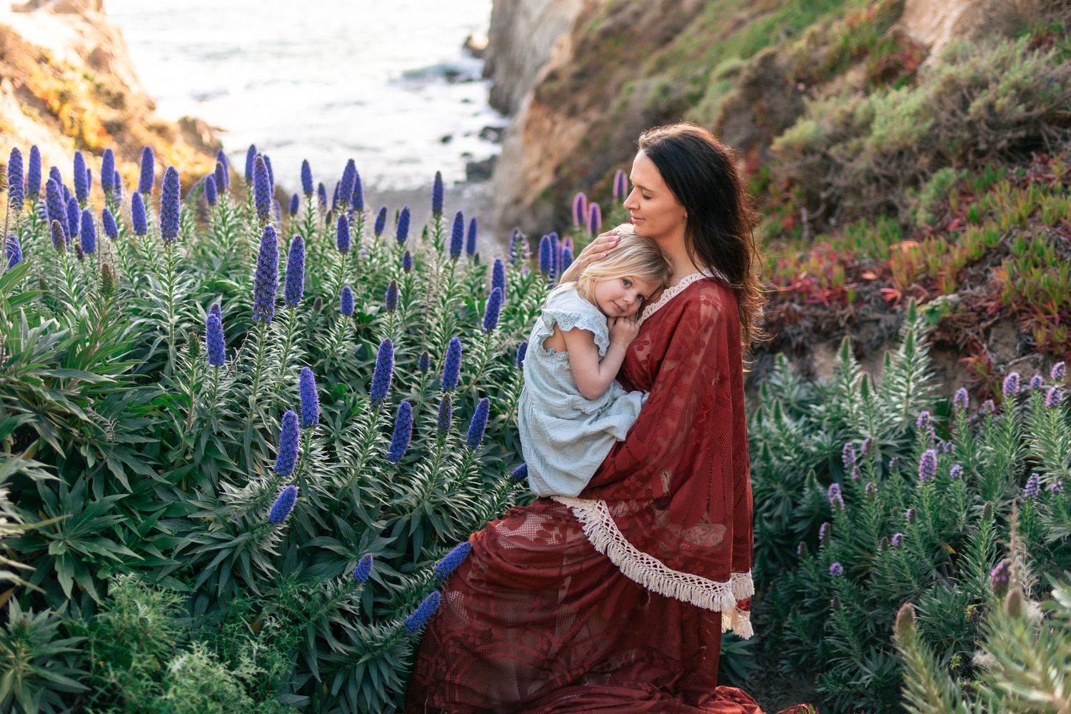 Woman in red dress holds young child among purple wildflowers on a scenic coastal cliffside.