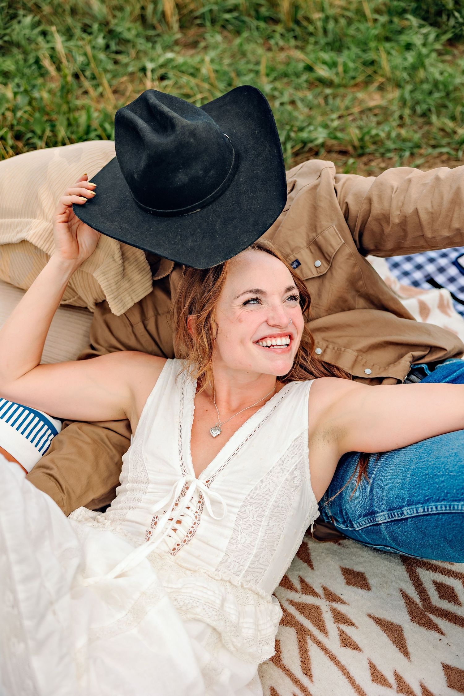 Smiling woman in white dress and jeans lying on blanket holding black cowboy hat outdoors.
