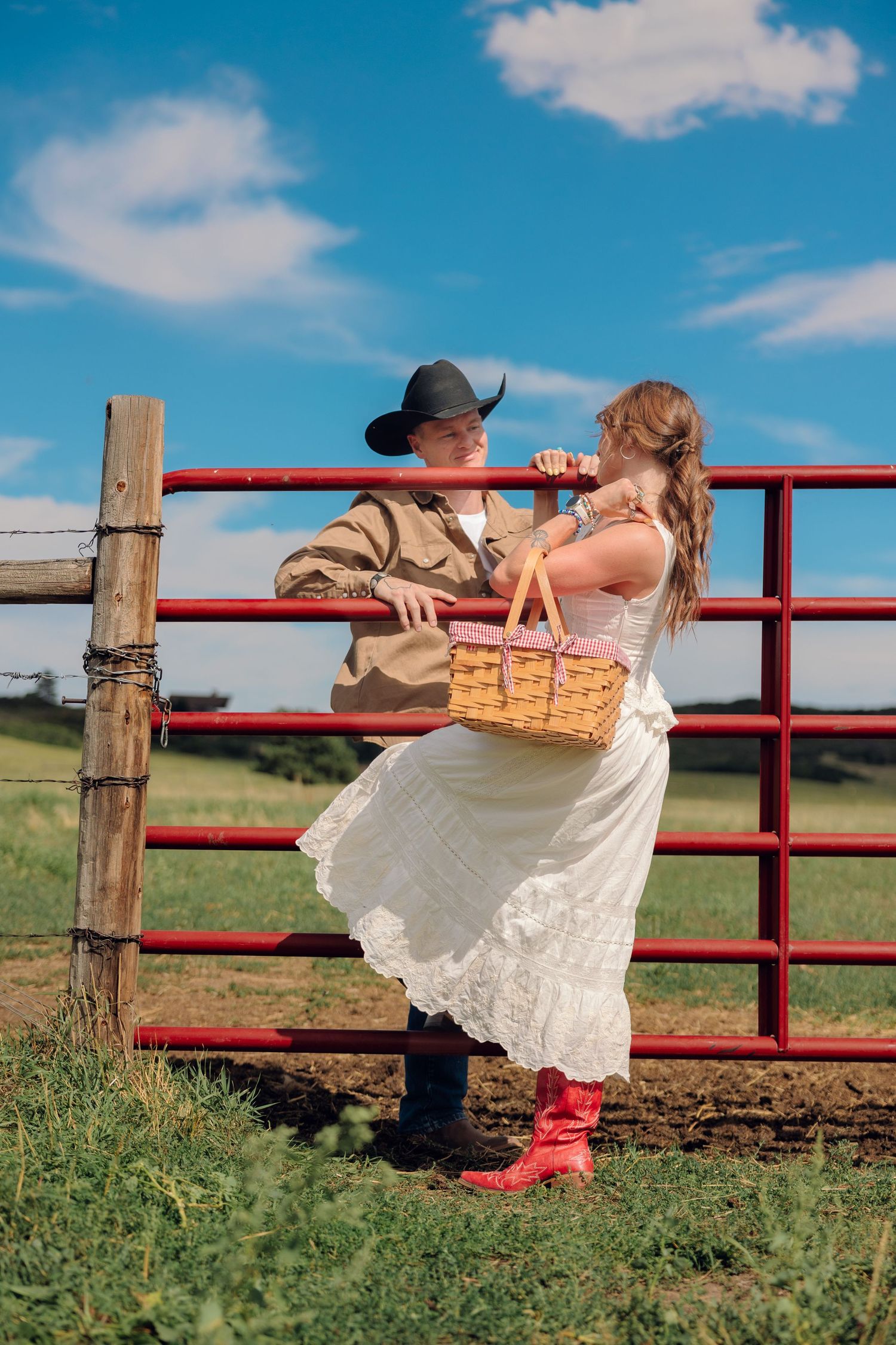 Couple in western attire by red farm gate, man in cowboy hat, woman in white dress with red boots holding a wicker basket.