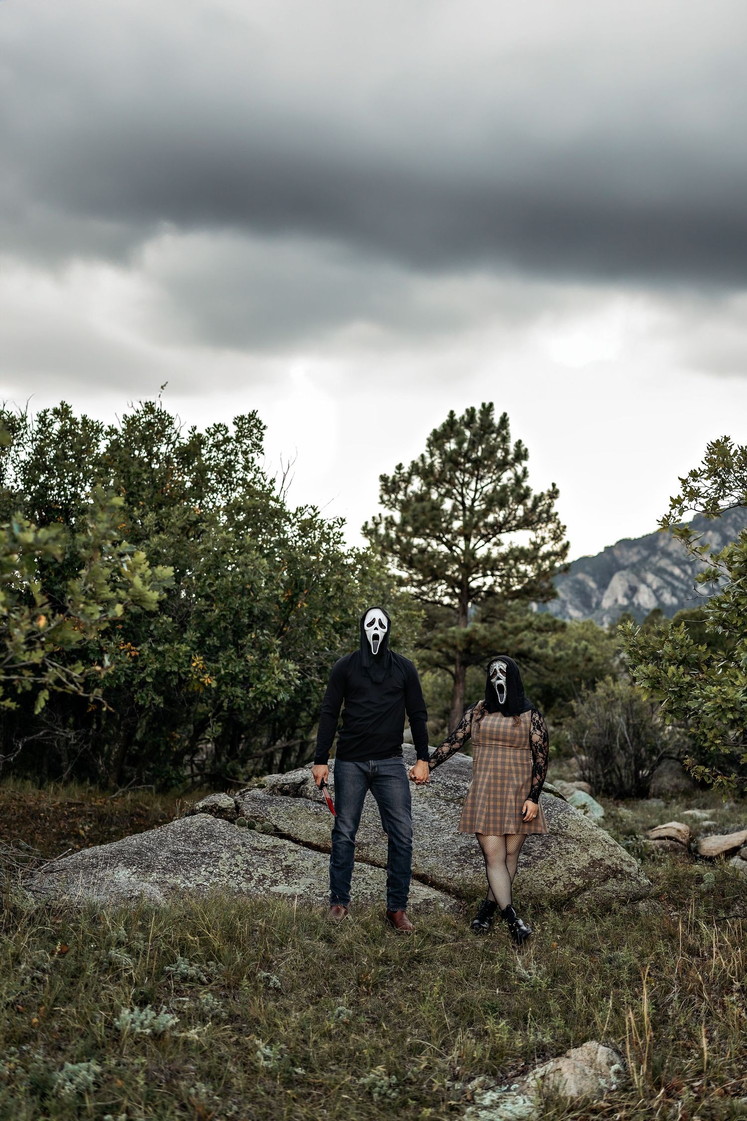 Couple in Ghostface masks holding hands on a large rock under a stormy gray sky, with pine trees and mountains behind.