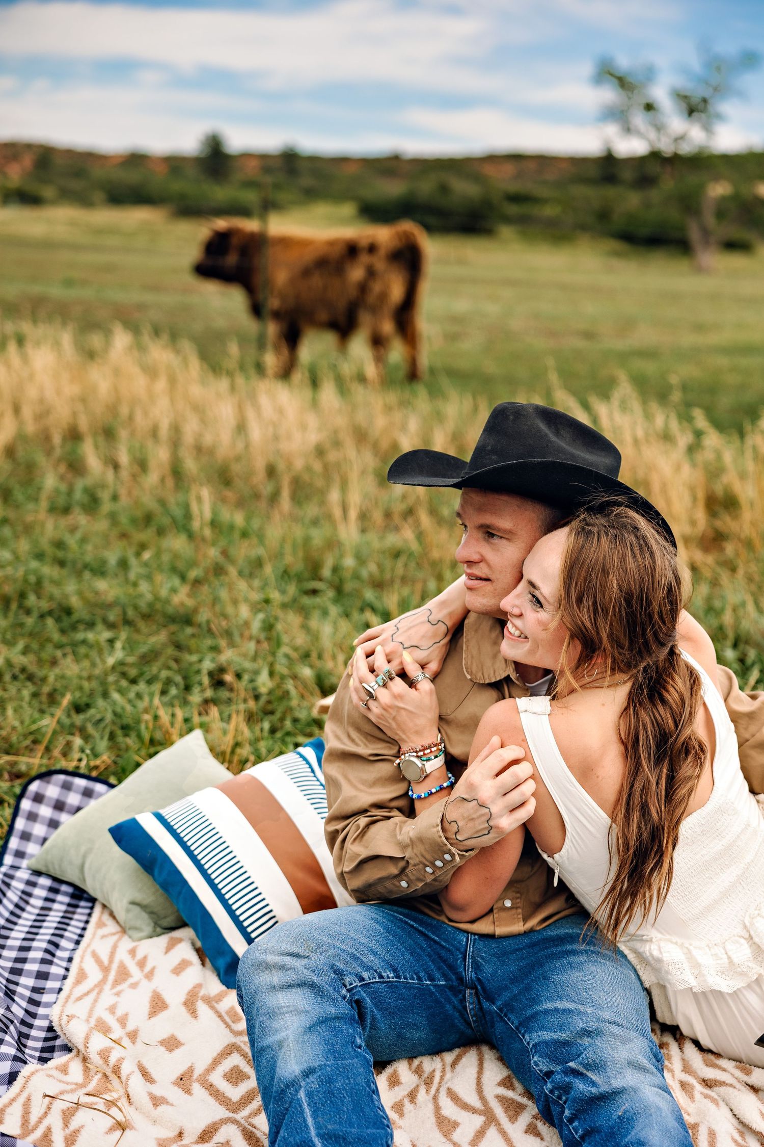 Couple sitting on a blanket in a field, man wearing cowboy hat, woman in white dress, with a cow grazing behind them.