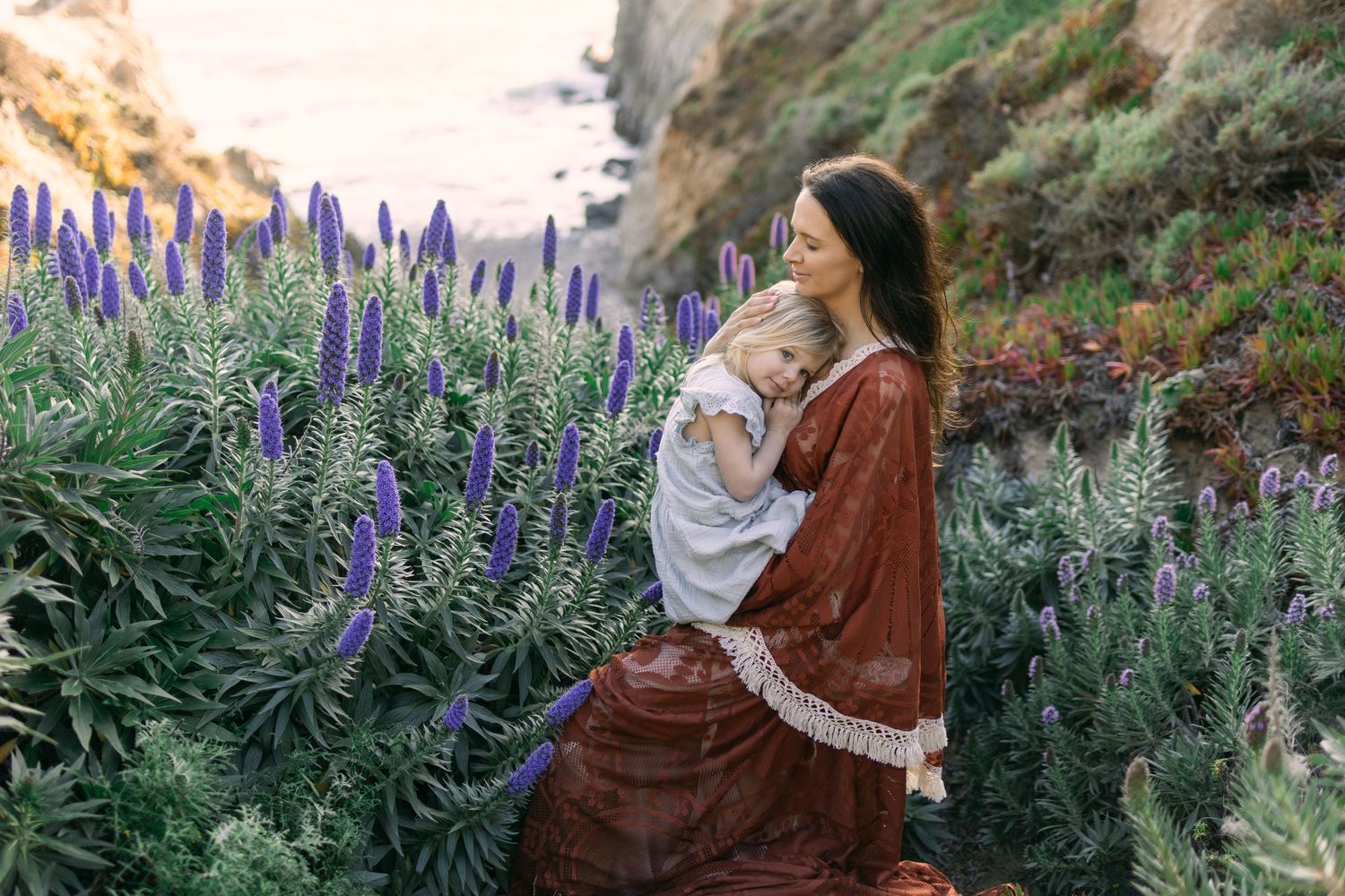 Mother holding baby among purple wildflowers on coastal cliffside, wearing bohemian red dress at golden hour.