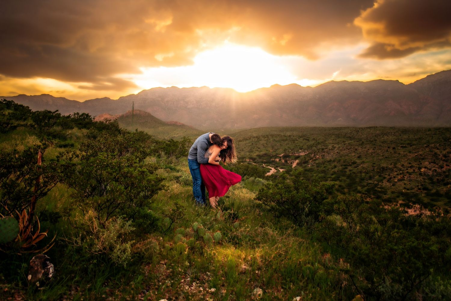 Couple embracing at golden sunset with dramatic mountain backdrop and lush green desert landscape.