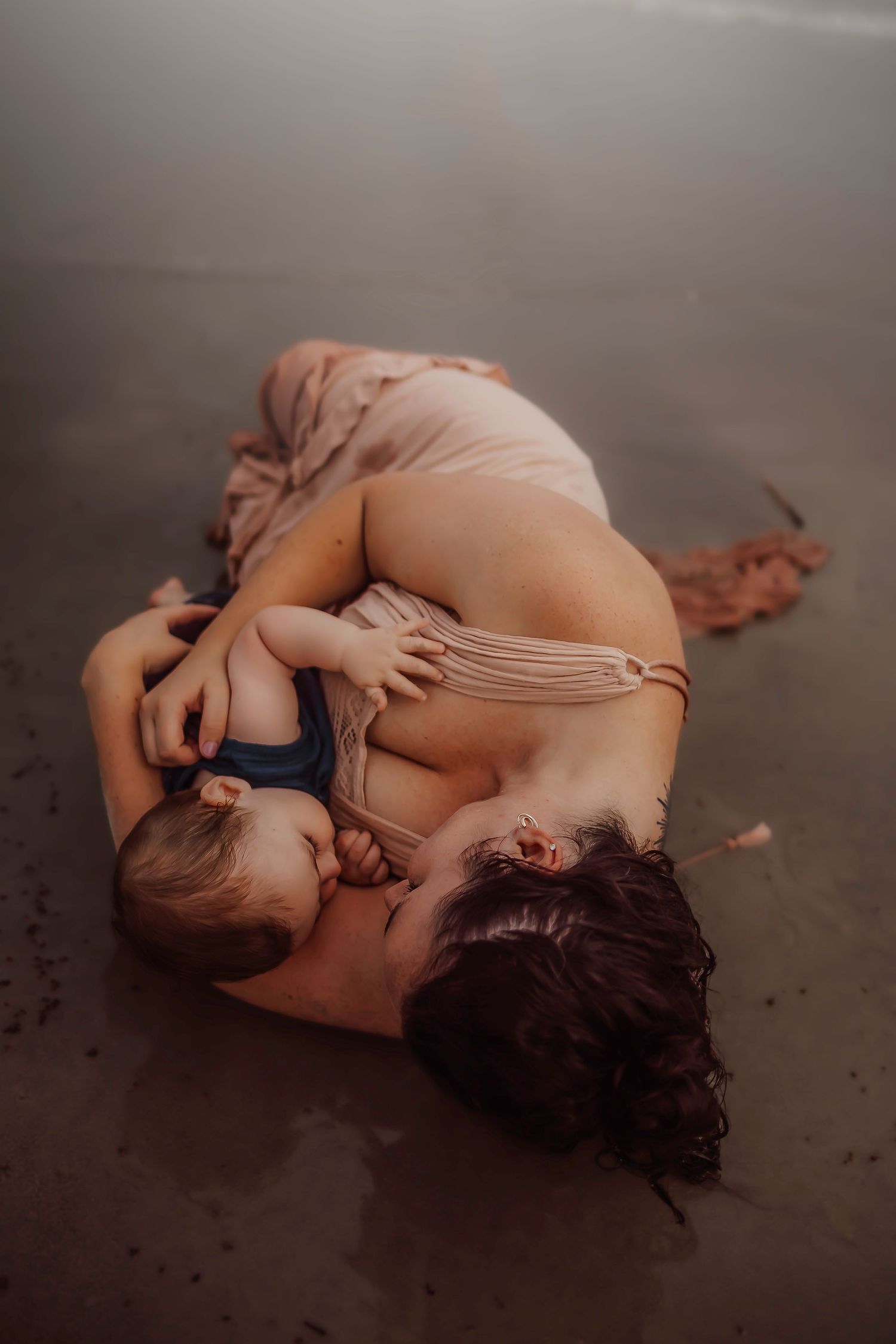 Mother lying on beach cradling and breastfeeding her infant baby in a tender, intimate moment.
