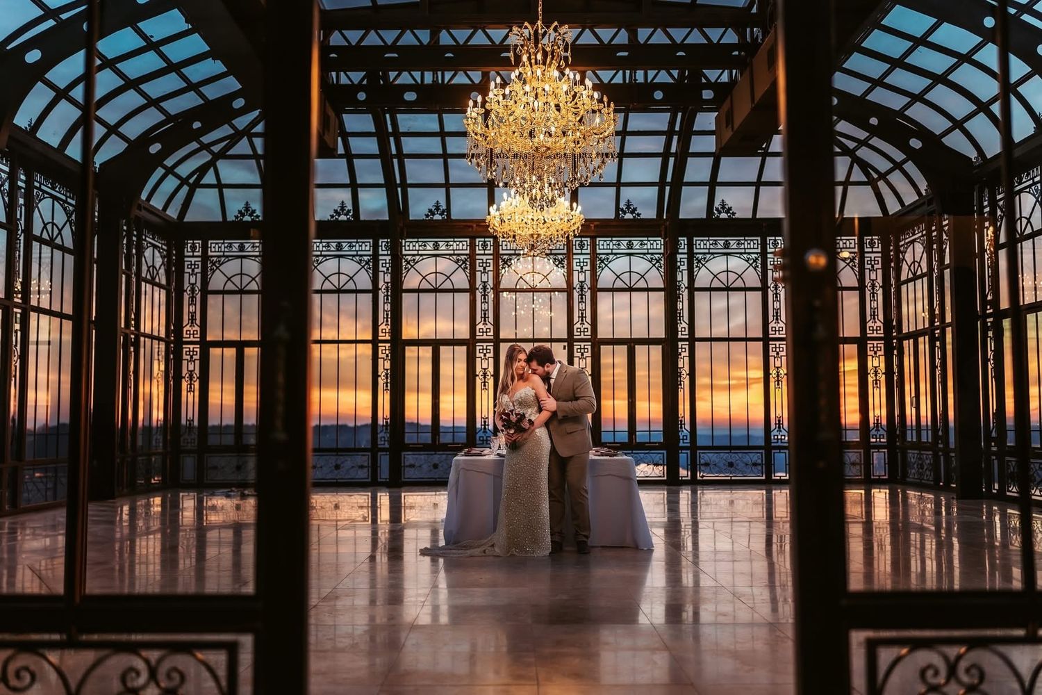 Couple shares a romantic kiss under a crystal chandelier in an elegant glass venue at sunset.