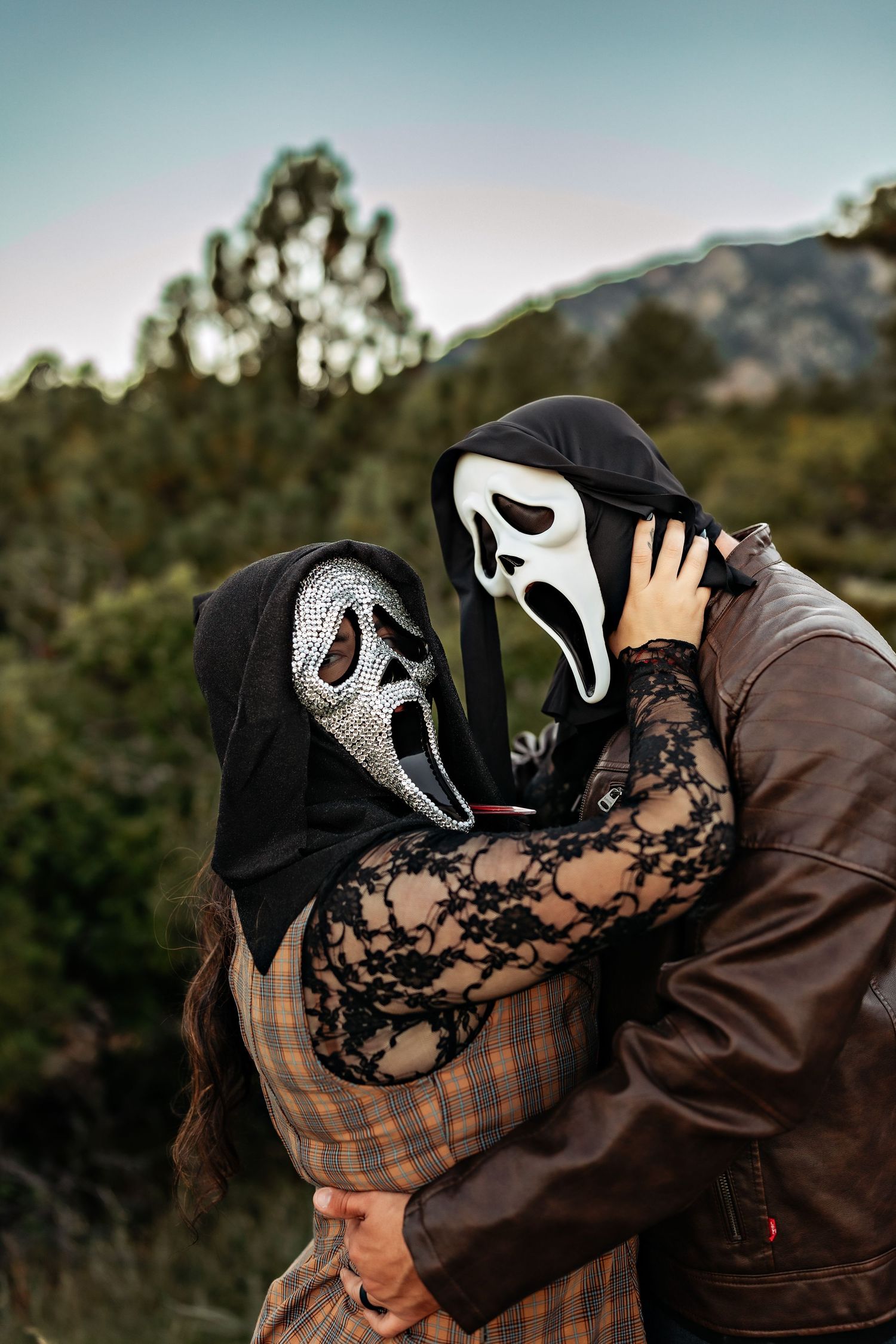 Couple embracing outdoors in Ghostface-style masks, one with a rhinestone mask and lace sleeves, before a mountain and trees at dusk.