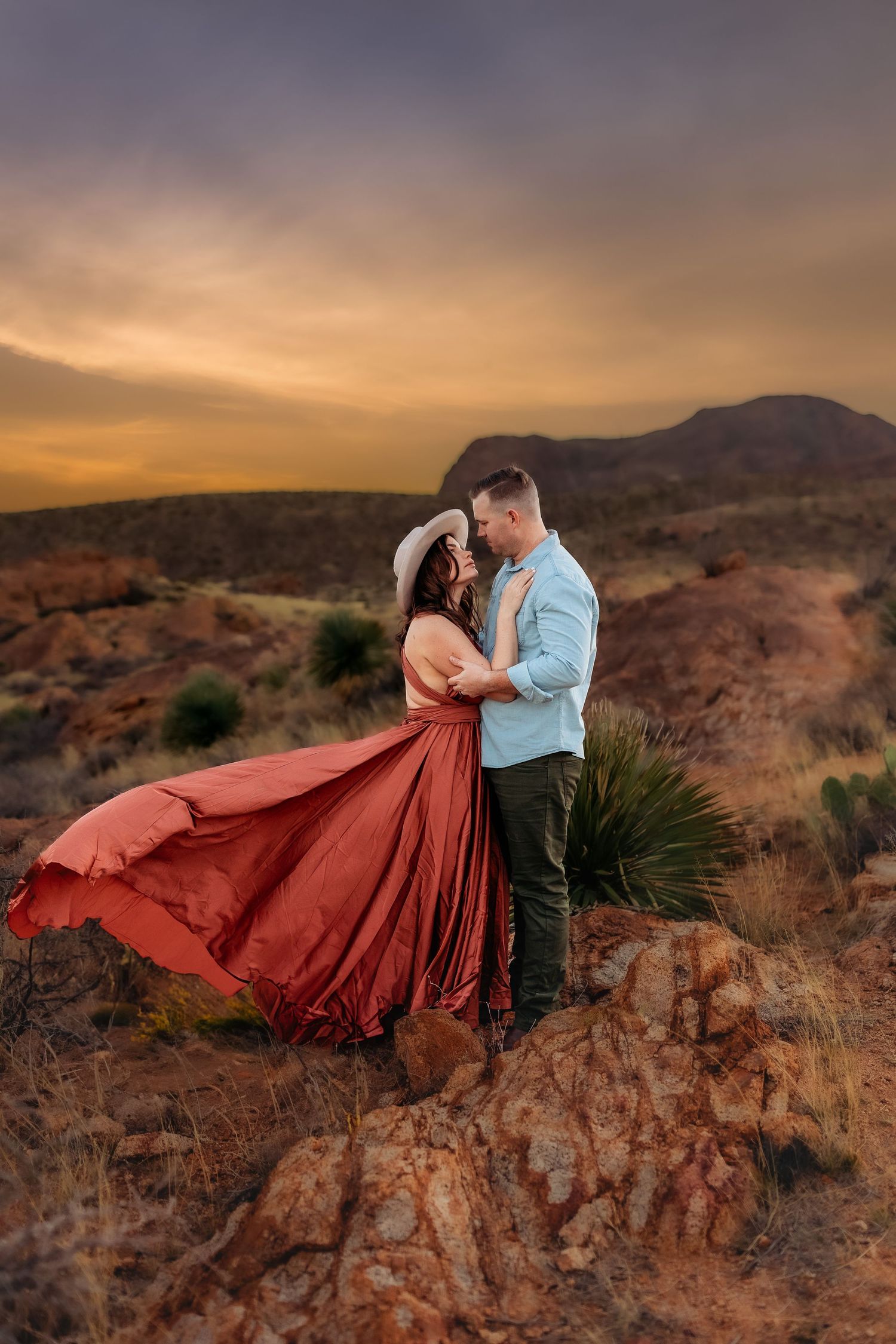 Couple embracing at sunset in red desert landscape, woman in flowing red gown and white hat, romantic golden sky.