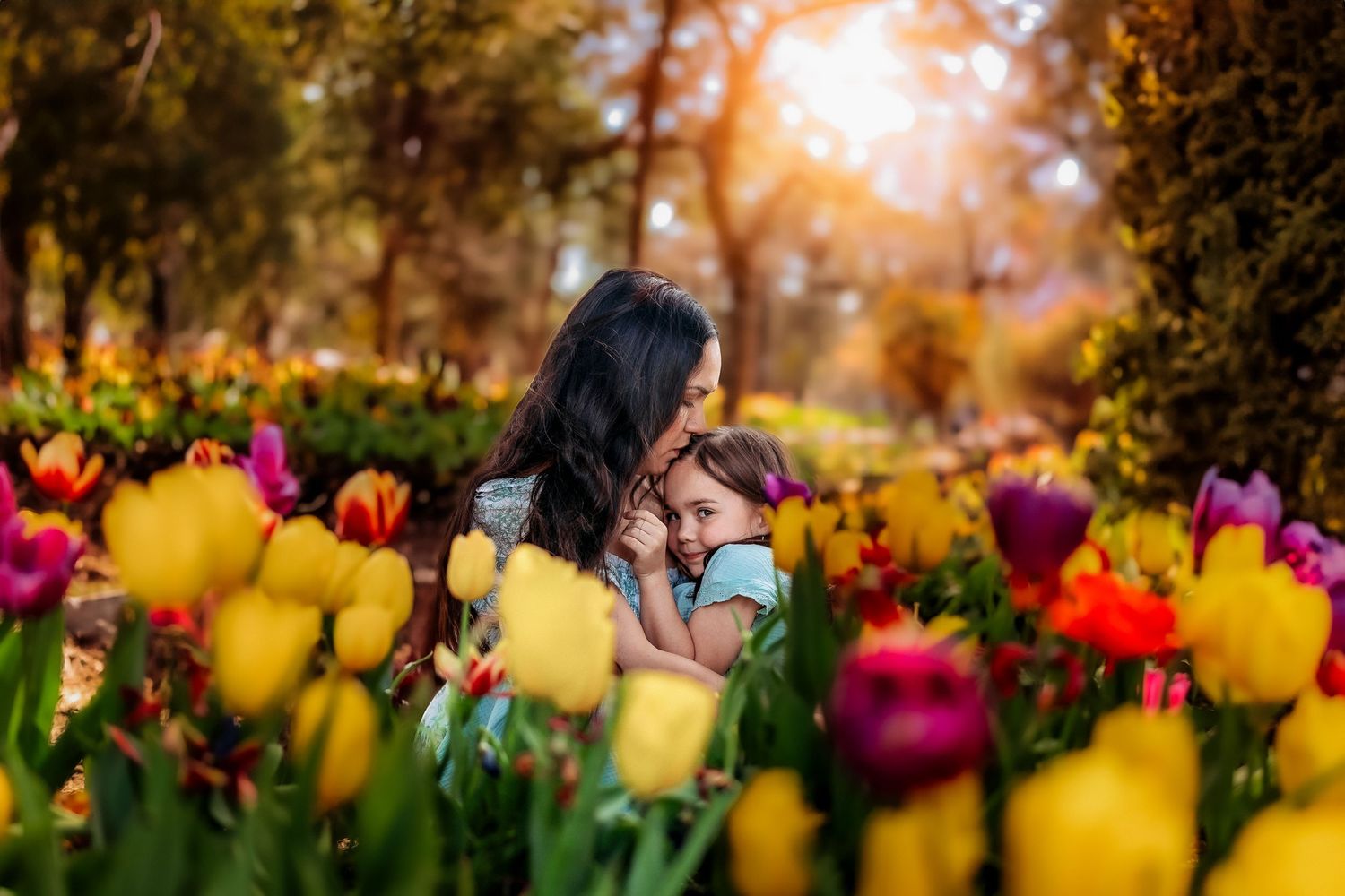 Mother and child embracing in a vibrant tulip garden with yellow, red, and purple flowers in warm sunlight.