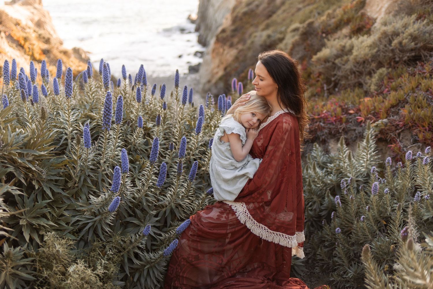 Mother in red dress holds baby among purple wildflowers on a scenic coastal cliffside.