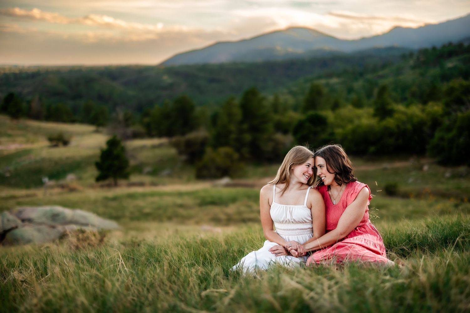 Two women in dresses sit together in a mountain meadow at sunset, sharing an intimate moment.