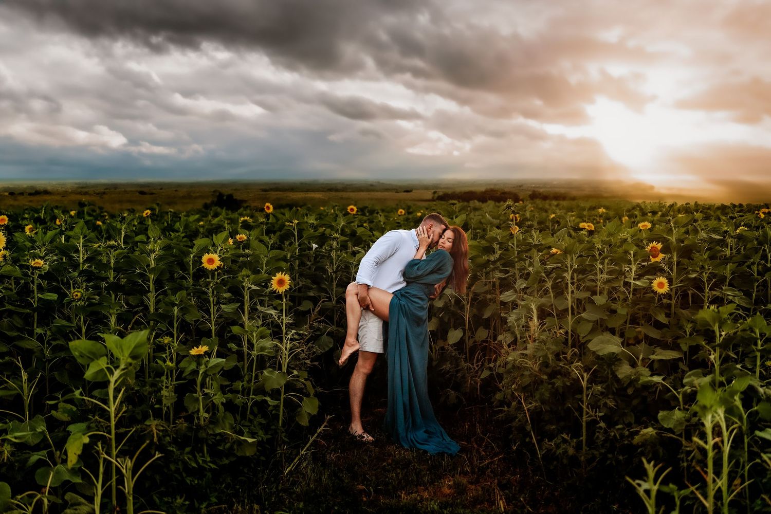 Couple embracing romantically in a sunflower field at golden sunset under dramatic stormy skies.