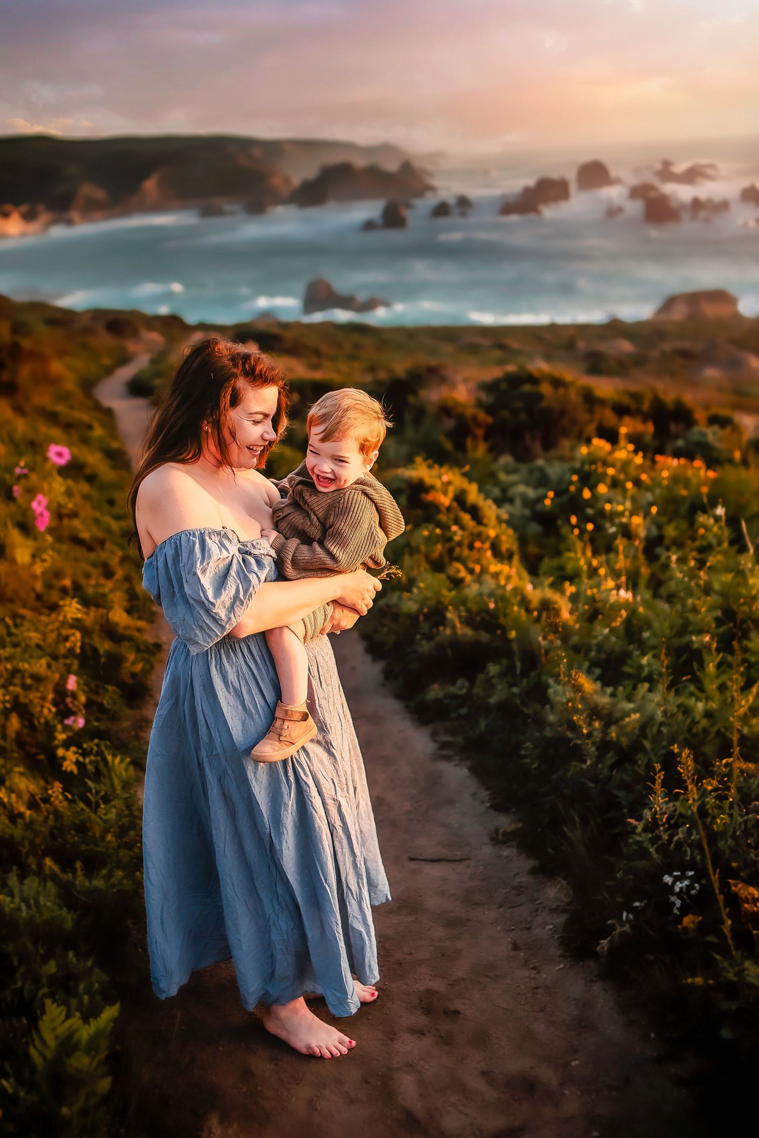 A woman in a blue dress holds a laughing toddler on a coastal wildflower path at golden hour.