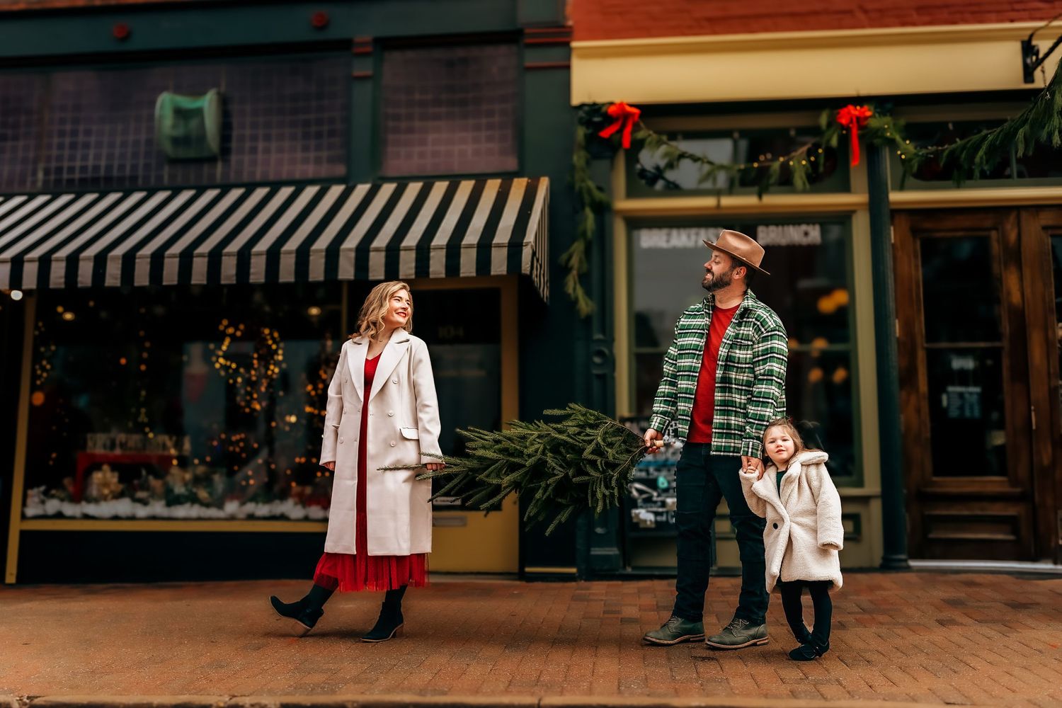 Family of three carrying a Christmas tree while shopping along a festive brick storefront street.