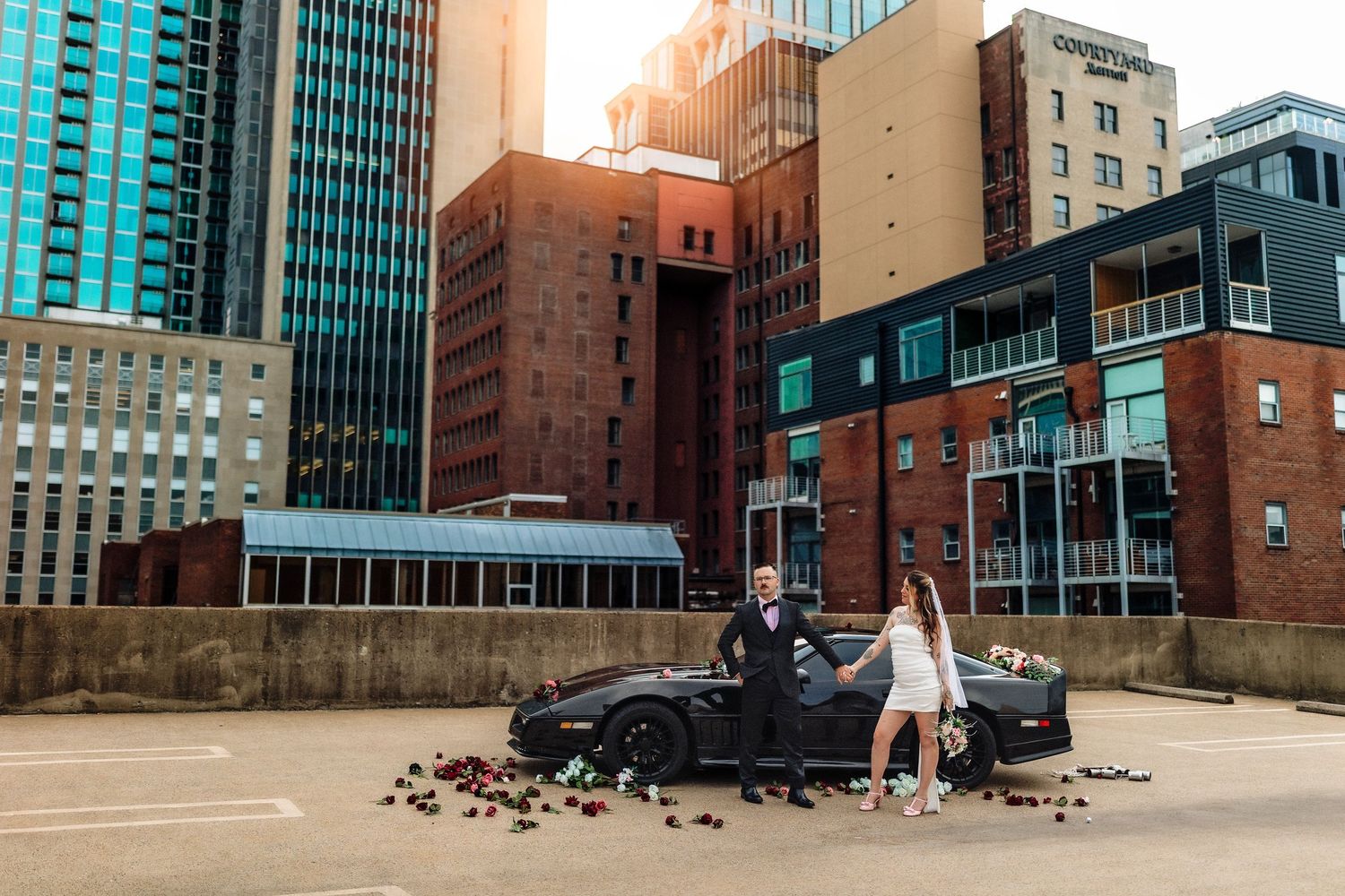 Couple poses by black sports car with rose petals scattered on ground, city skyline with brick buildings in background.