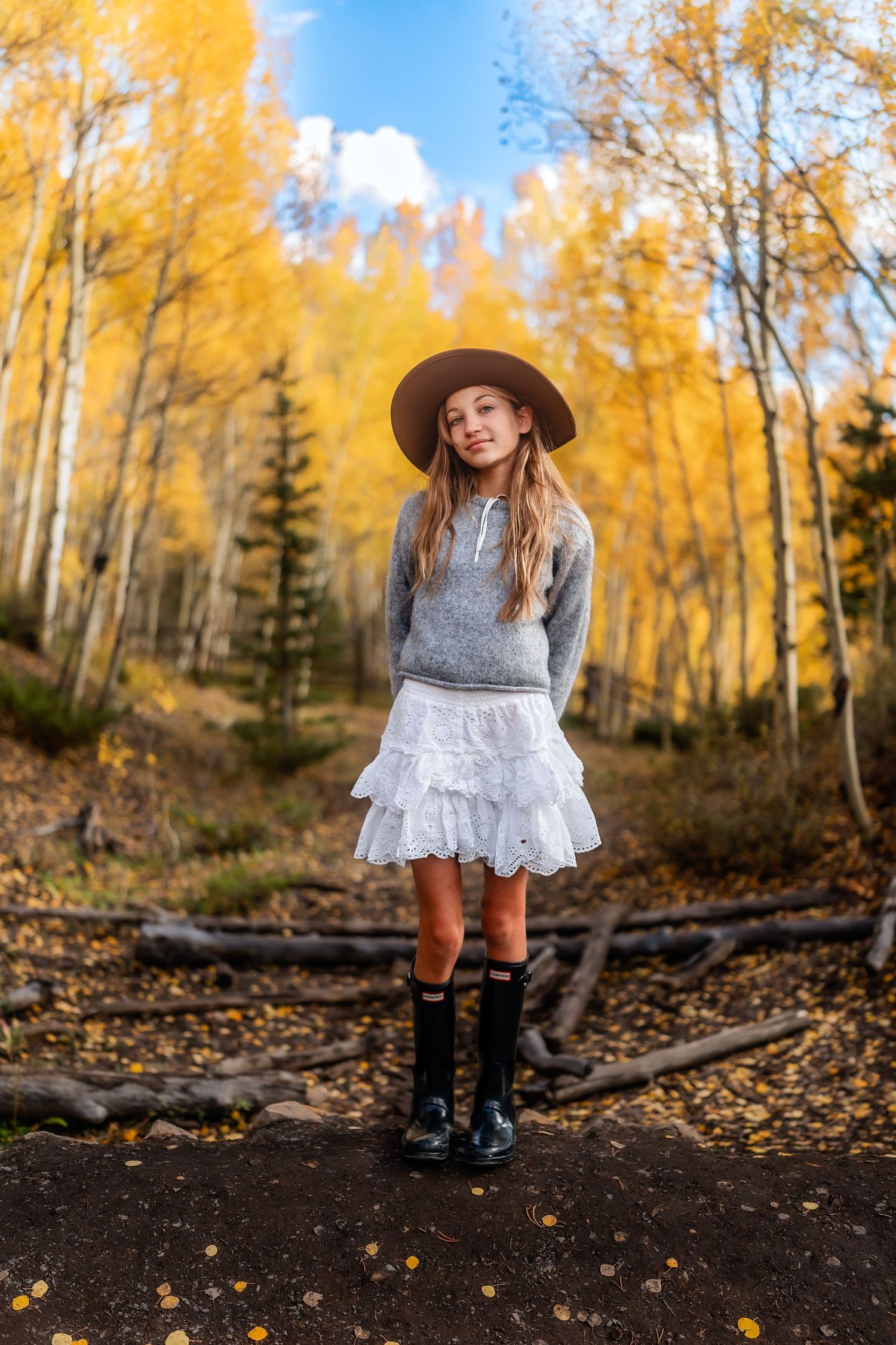 A young girl in a gray sweater, white ruffle skirt, brown hat, and black boots stands among golden autumn aspen trees.