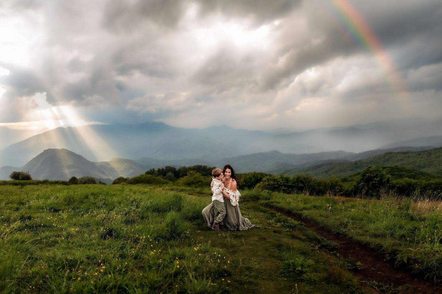 Mother and child embracing on a green mountain meadow with a rainbow and dramatic cloudy sky in the background.