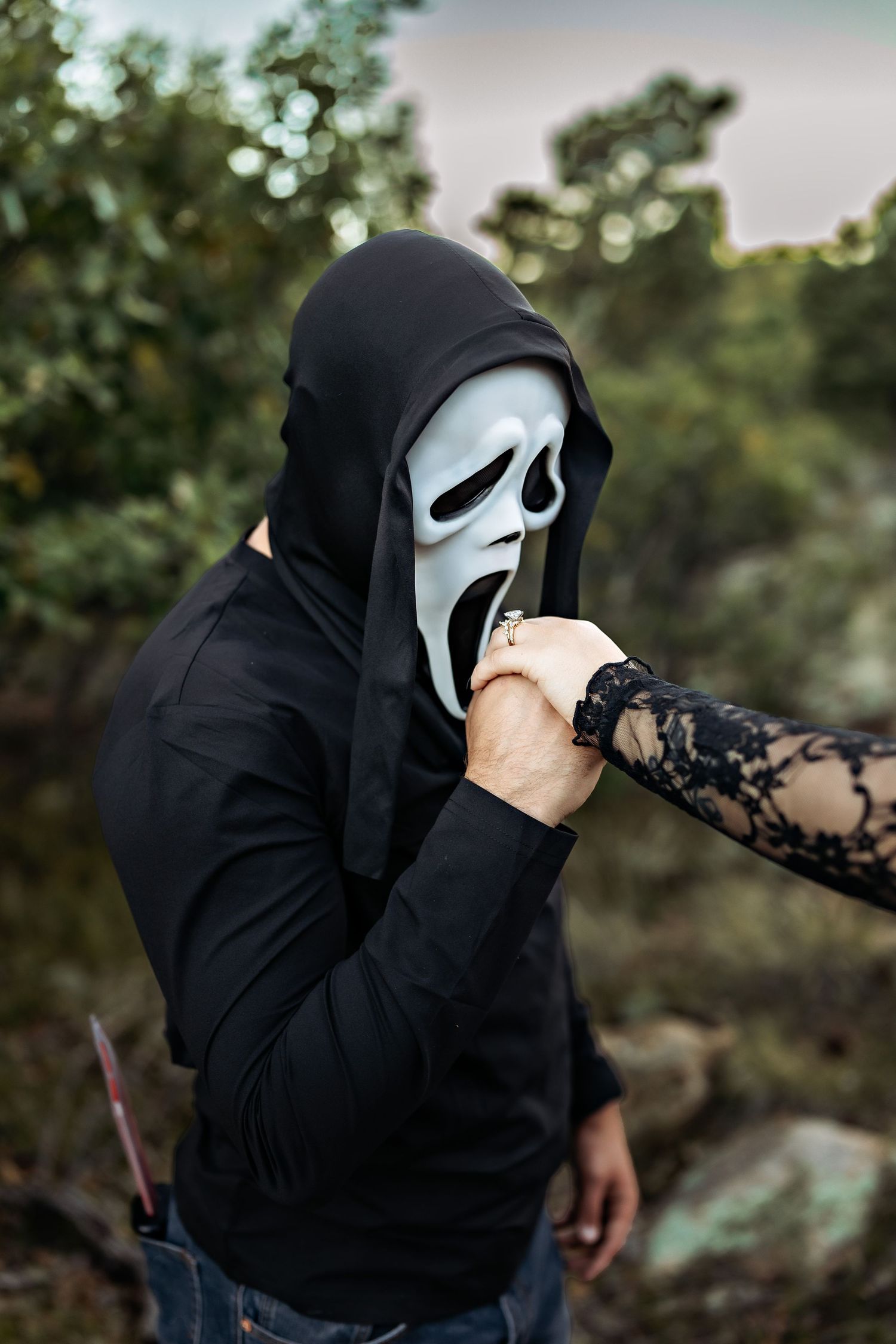 Person in a black hoodie and ghost face mask holds a knife while a woman in a lace sleeve kisses their hand in a rugged outdoor setting.