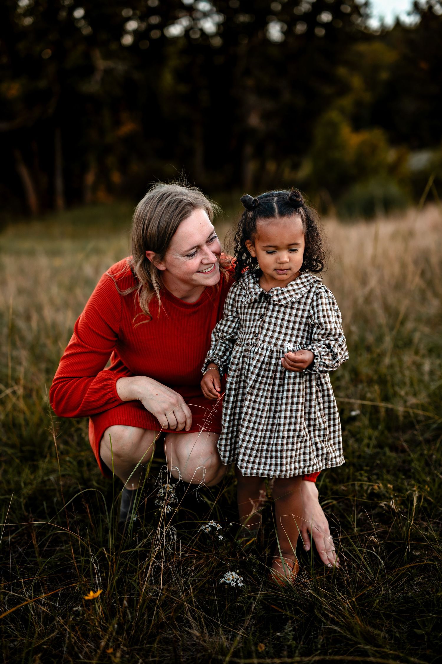 A smiling woman in a red dress kneels in a grassy field beside a young girl in a checkered dress at golden hour.