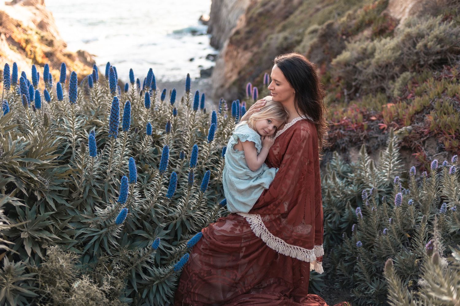 Mother in red dress holds baby among blue wildflowers on coastal cliffside at sunset.