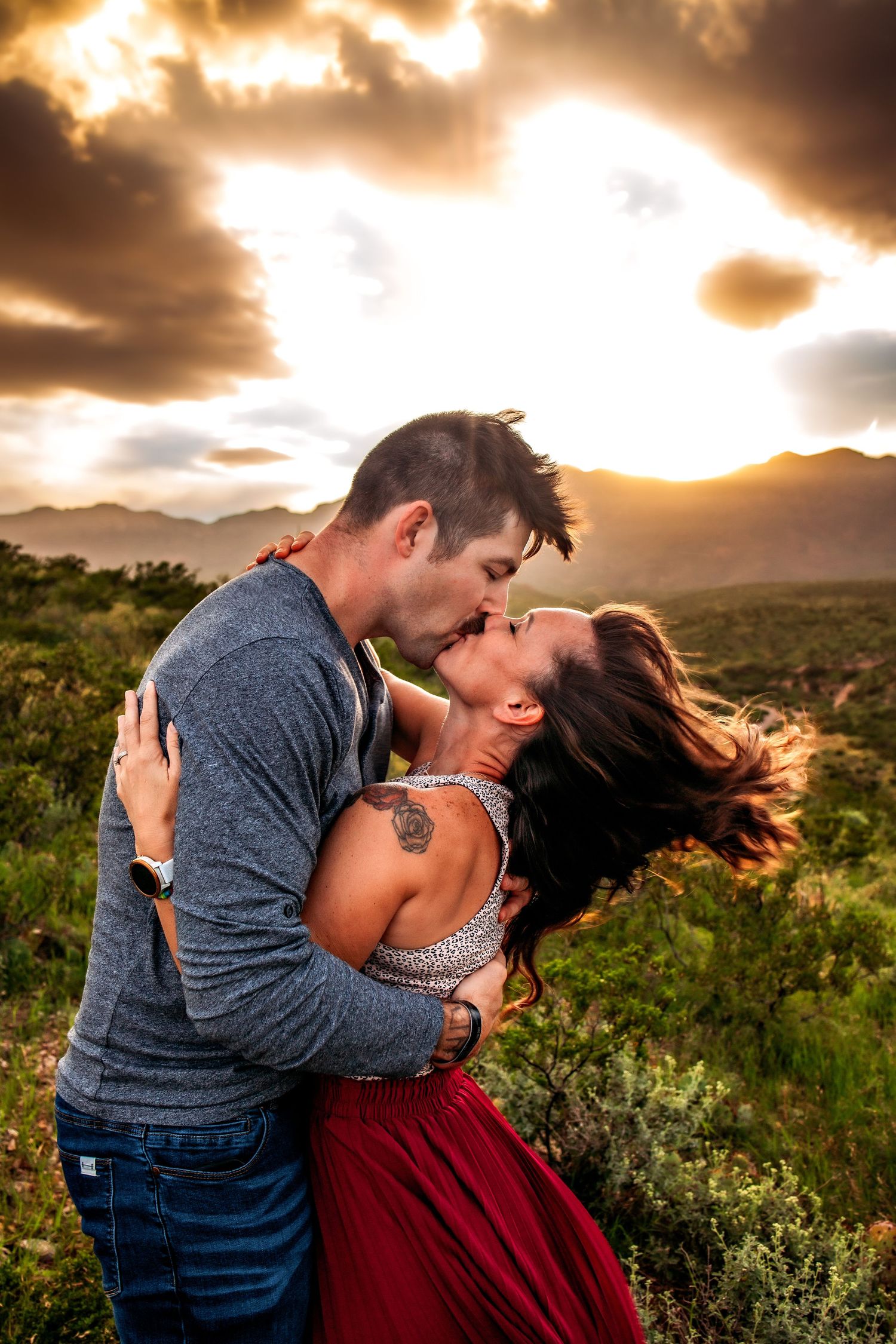 Couple sharing a passionate kiss at sunset with dramatic clouds and mountain landscape in the background.