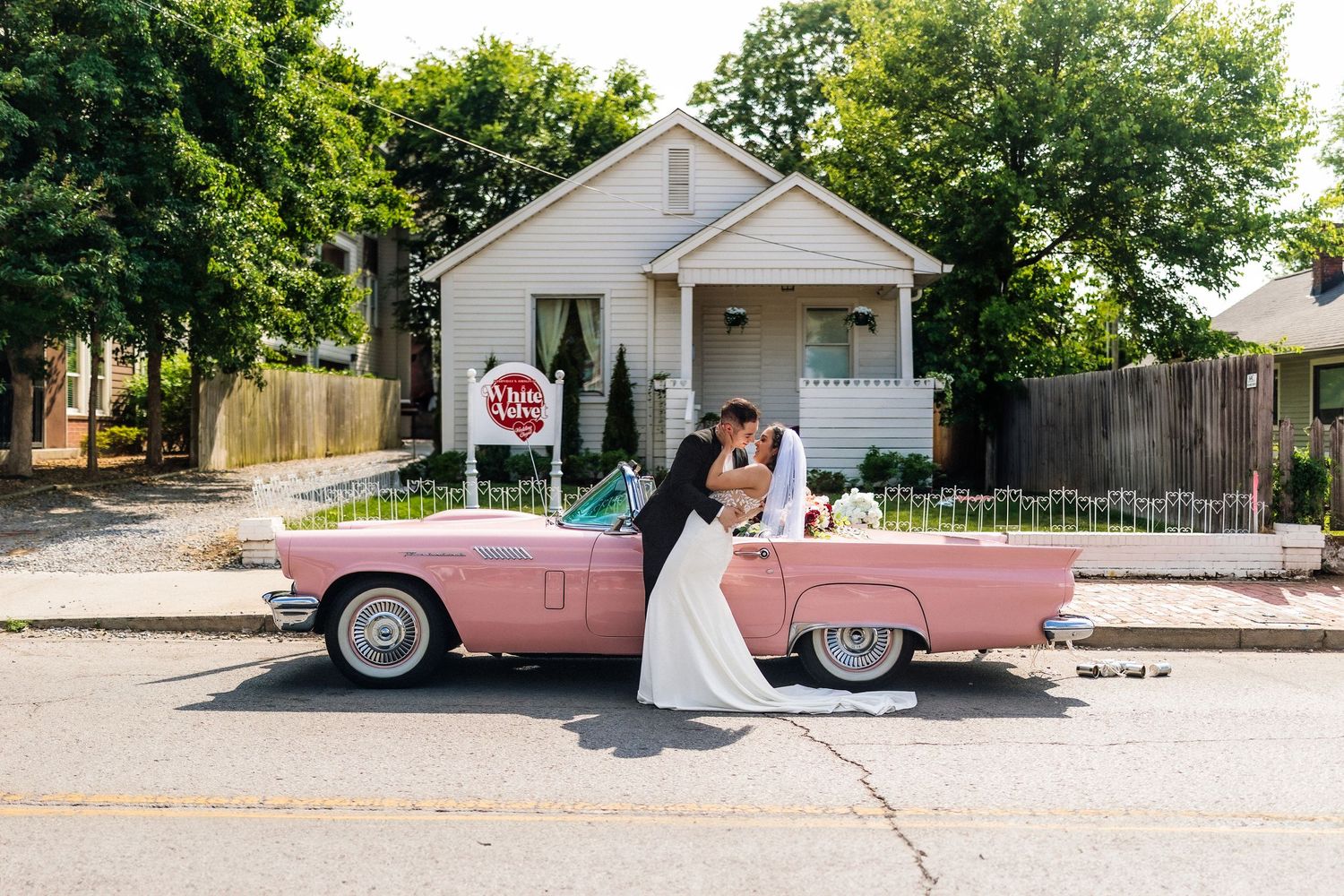 Bride and groom kissing beside a classic pink convertible car on a sunny street with a white house in the background.