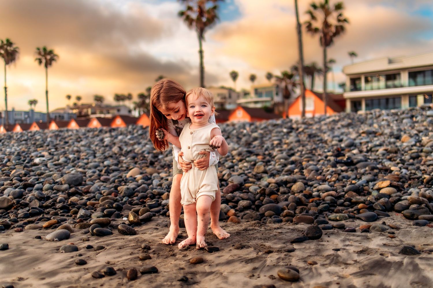 Mother helping smiling baby stand on rocky beach at sunset with palm trees and colorful sky in background.