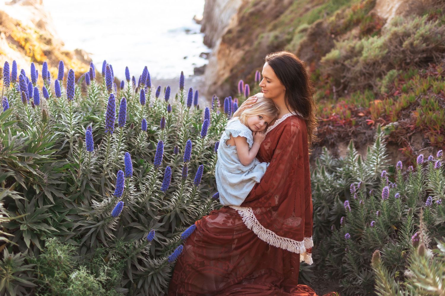 Mother in red dress holds young child among purple wildflowers on a coastal cliffside.