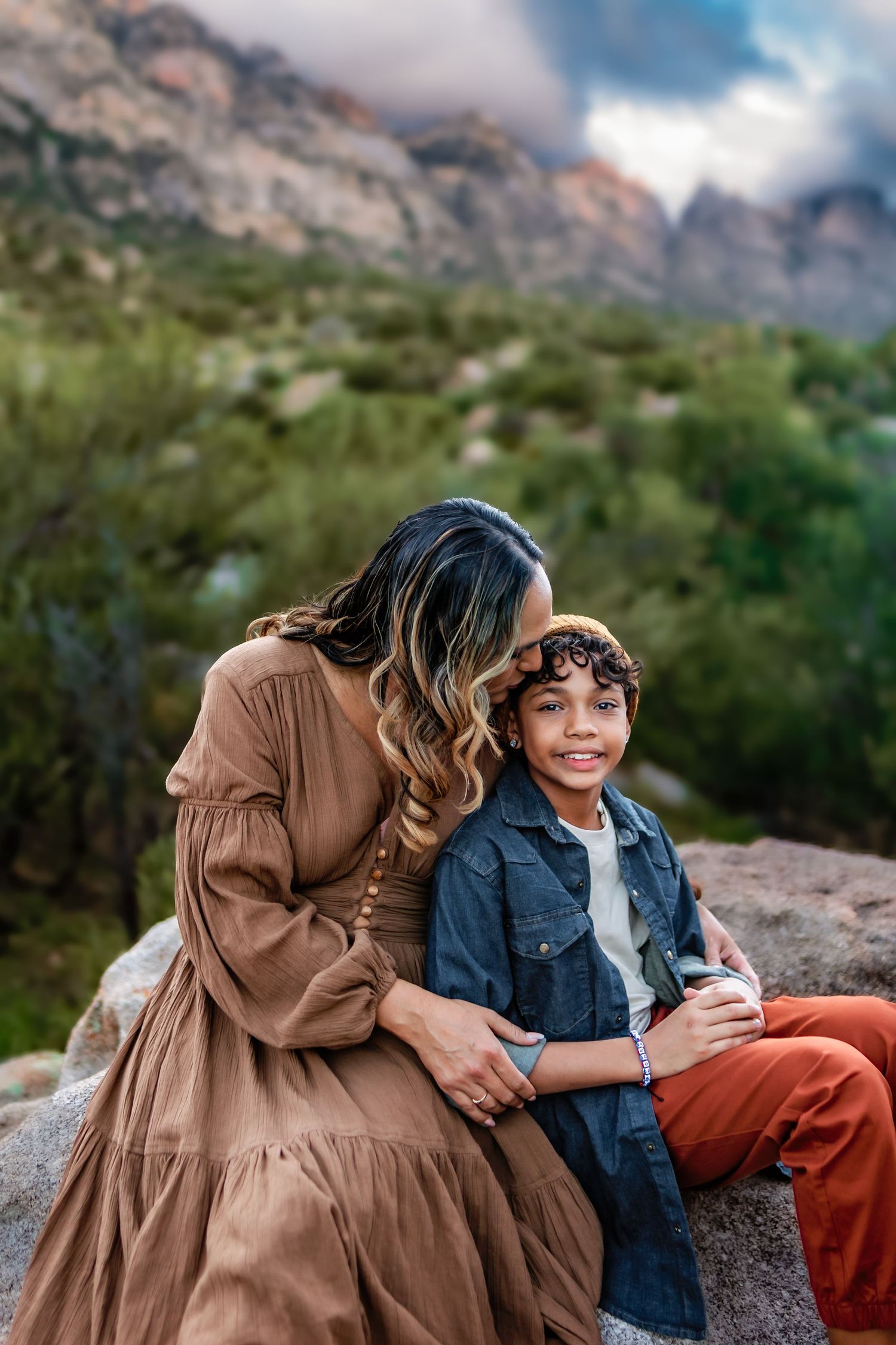 A mother in a brown dress hugs her smiling son on a rocky outcrop with mountains and greenery behind them.